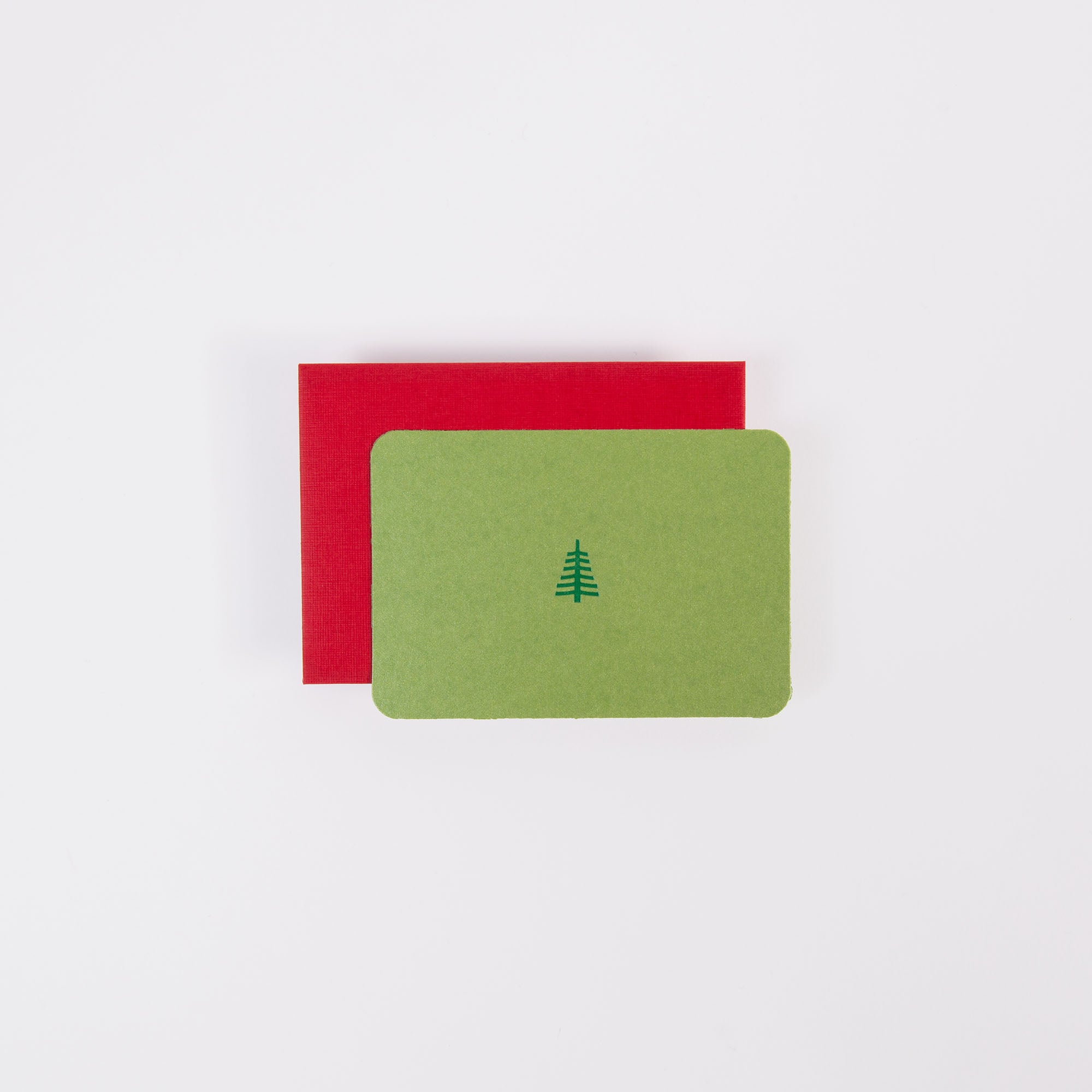 Die Mini Karte "Tannenbaum klein" von Le Typographe zeigt ein schlichtes Tannenbaum-Icon auf grünem Karton und liegt auf einem roten Umschlag - ein perfektes Weihnachtsmotiv vor weißem Hintergrund.