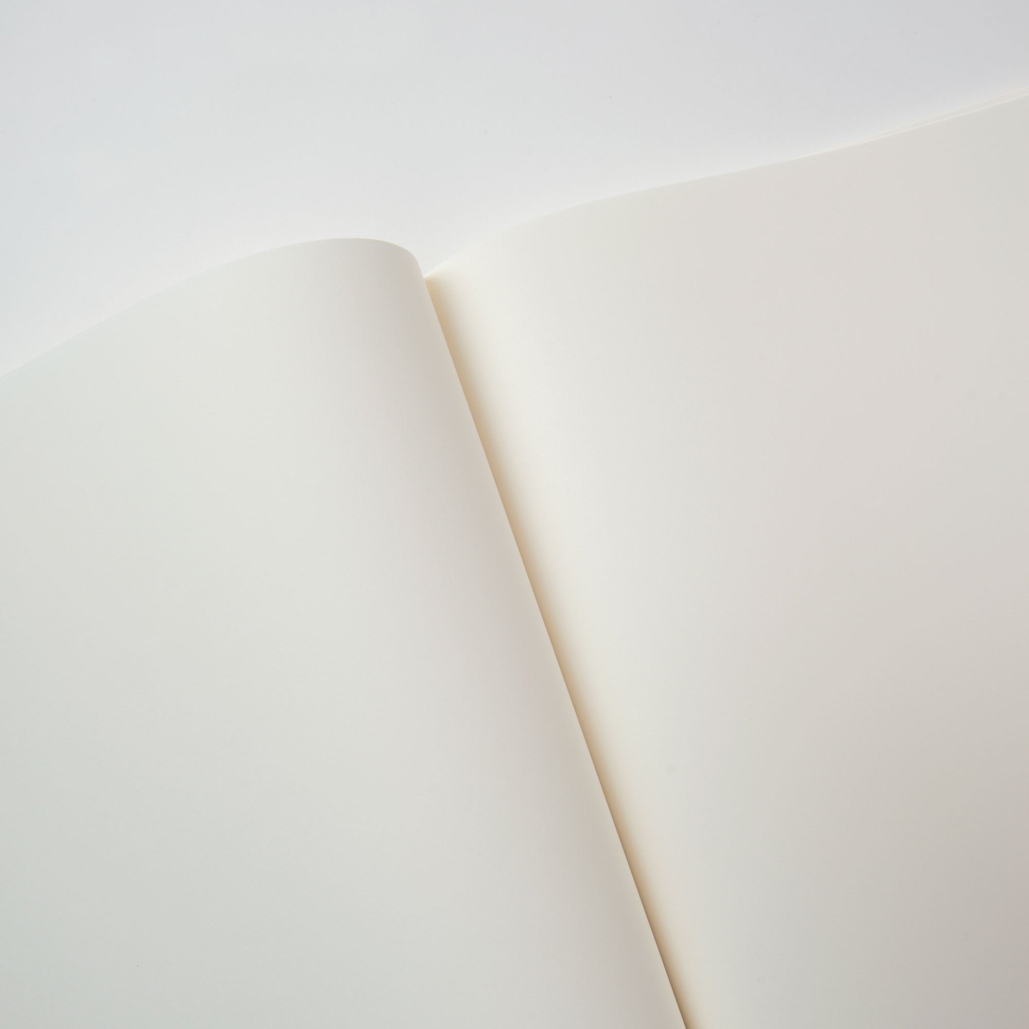 Bei sanfter Beleuchtung zeigt ein Notizheft mit Format von Midori cremeweiße Seiten aus MD Cotton Feinstpapier. Die Fadenbindung erzeugt eine sanfte Kurve in der Nähe des Rückens, wirft zarte Schatten und hebt die minimalistische Textur und Reinheit des Papiers hervor.
