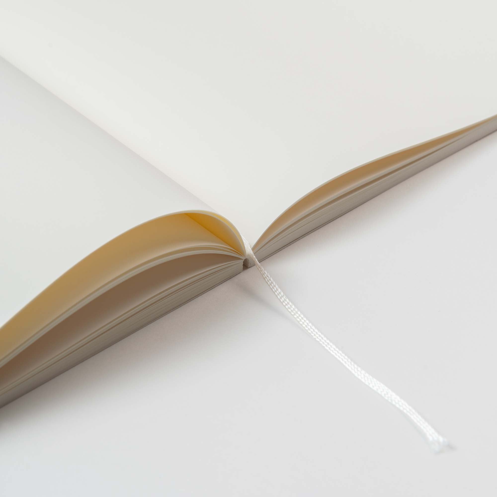 Nahaufnahme eines offenen Midori Ein Notizhefts mit Format mit leeren, cremefarbenen MD Cotton Feinstpapier-Seiten und einem weißen Lesezeichenband. Auf einer glatten weißen Oberfläche platziert, strahlt die Szene eine minimalistische und klare Ästhetik aus.