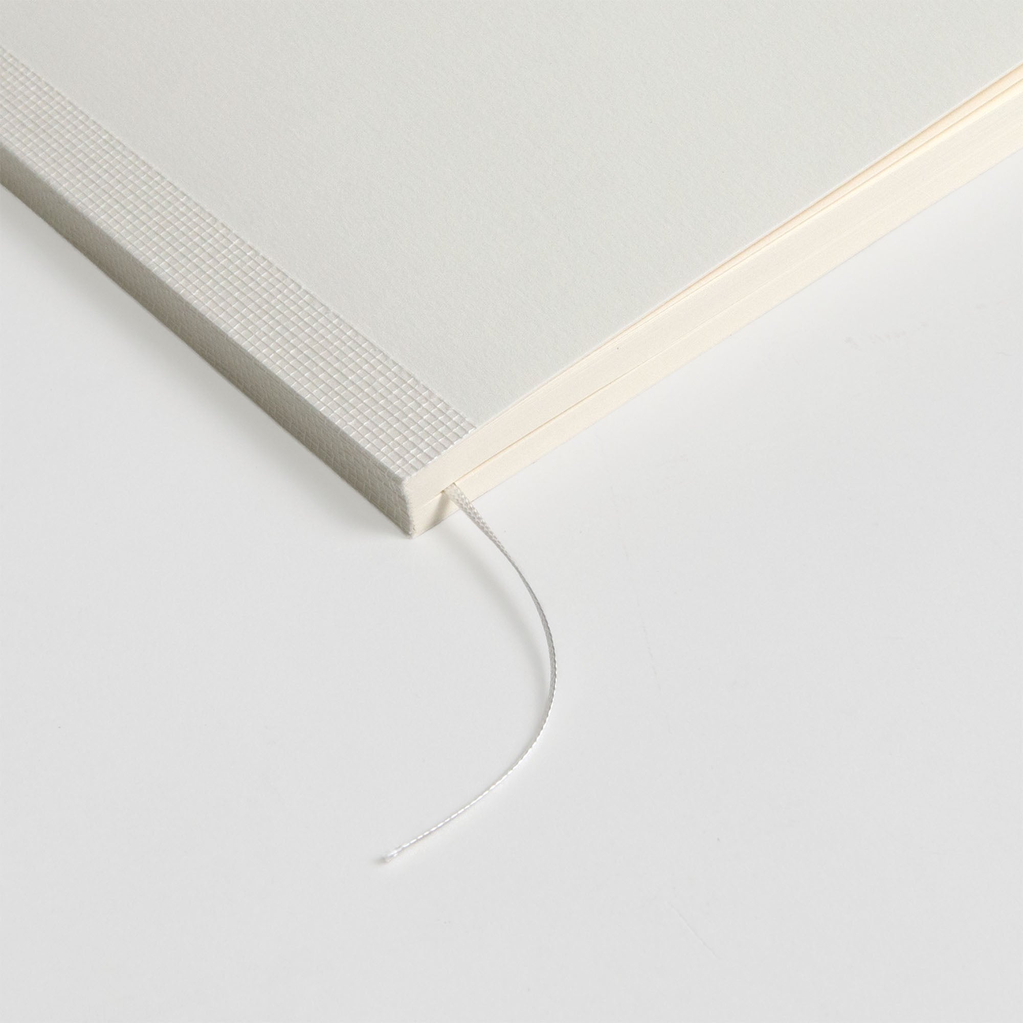 Eine Nahaufnahme der Ecke eines weißen Midori MD Notizbuchs A5 Baumwolle mit strukturiertem Rücken und Fadenheftung, mit einem dünnen weißen Lesezeichen auf einer weißen Oberfläche.