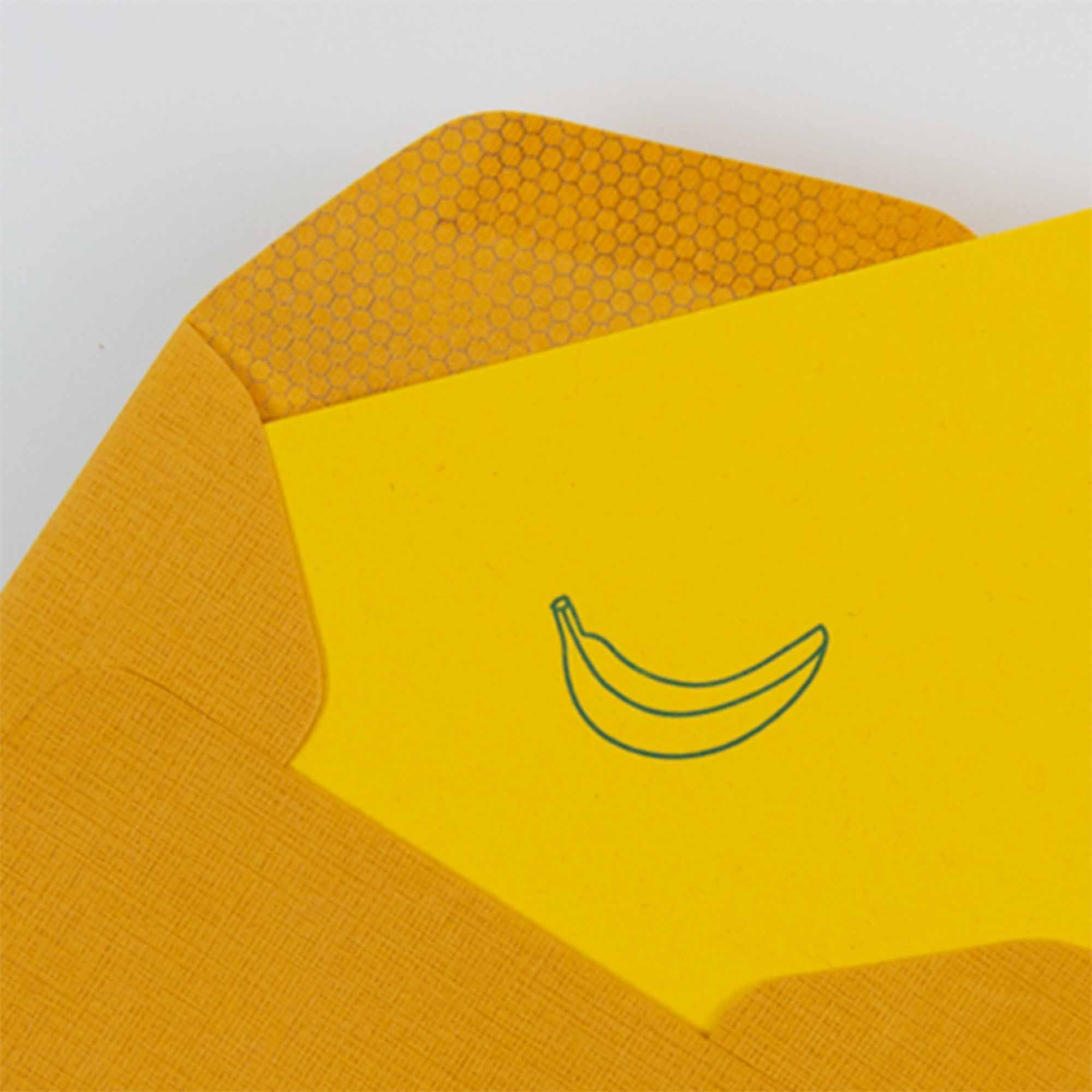 Eine Le Typographe „Banana - Mini Karte“ steckt in einem gelben Umschlag mit Wabenmuster. Die Karte zeigt eine grün umrandete Banane im Buchdruckverfahren, was die exquisite Handwerkskunst unterstreicht, die an traditionelle Buchbindereitechniken erinnert.