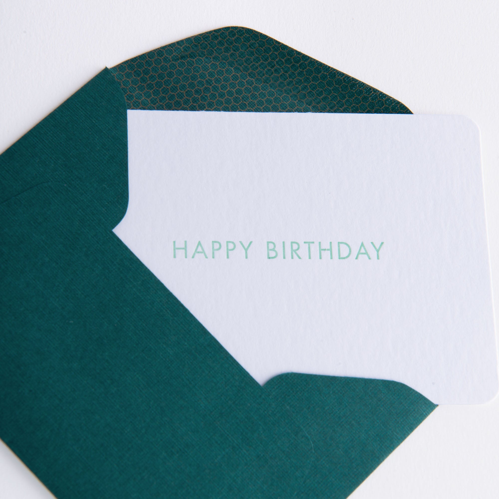 Eine weiße „Happy Birthday - Minikarte“ von Le Typographe mit hellblaugrünem Text ist teilweise aus einem dunkelblaugrünen Kuvert herausgezogen. Die innere Klappe des Umschlags weist ein subtiles geometrisches Muster auf, das an komplizierte Buchdrucktechniken erinnert.