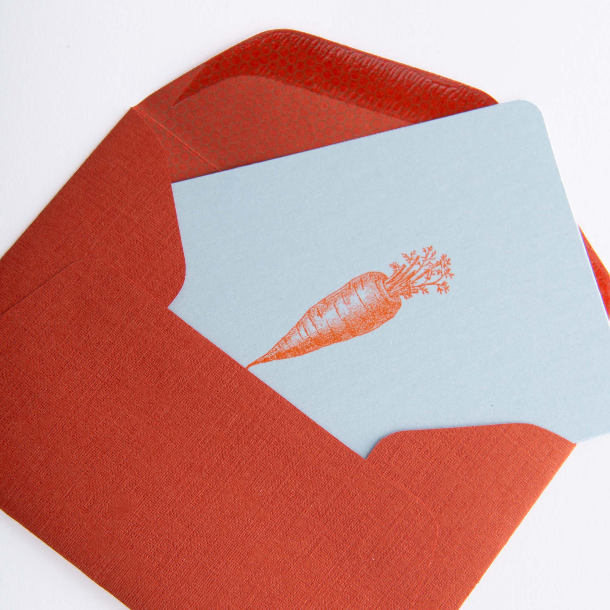 Eine hellblaue „Karotte Orange - Mini Karte“ von Le Typographe mit einer orangefarbenen Karottenillustration lugt aus einem orangefarbenen strukturierten Umschlag mit roter Innenseite hervor. Dieses elegante Karten- und Umschlagset wurde im Buchdruckverfahren hergestellt und hebt sich vom weißen Hintergrund ab.