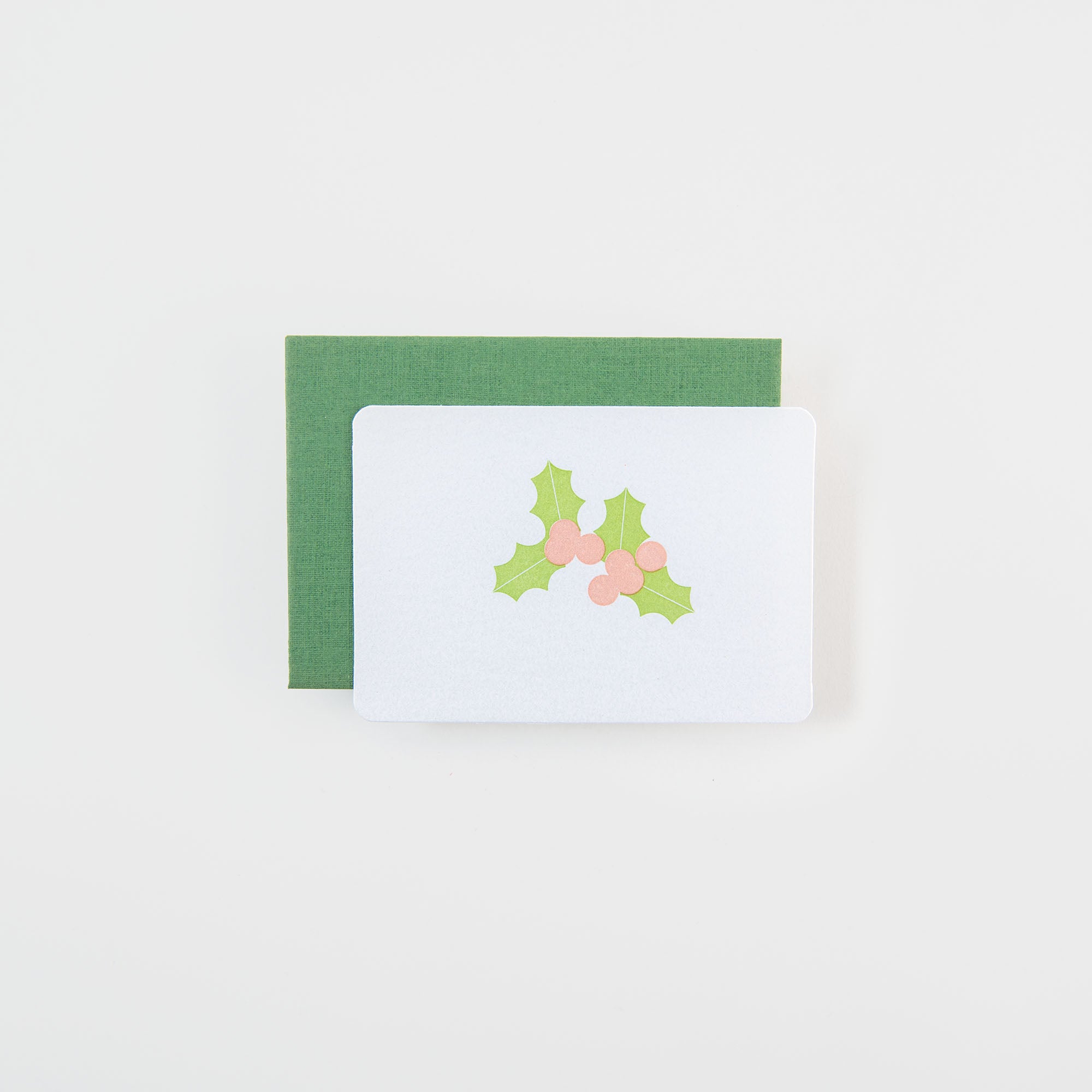 Die Mistelzweig - Mini Karte von Le Typographe ist eine weiße Weihnachtskarte mit grünen Stechpalmenblättern und roten Beeren, die auf einem grünen Umschlag vor einem schlichten weißen Hintergrund abgebildet ist.