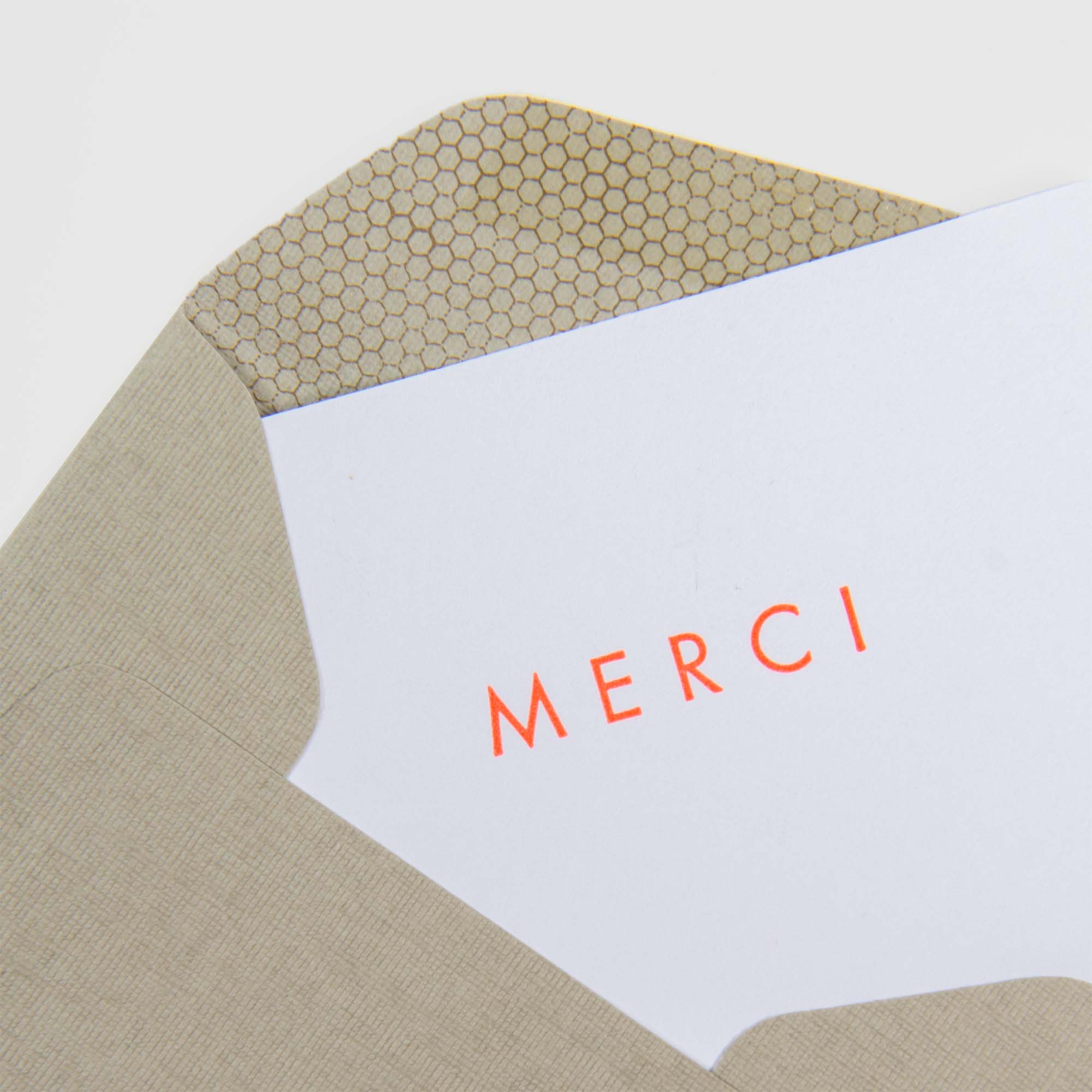 Die Merci - Mini Karte von Le Typographe ist eine weiße Letterpress-Karte mit dem roten Schriftzug "MERCI", die in einem beigen Umschlag mit Wabenmuster steckt.