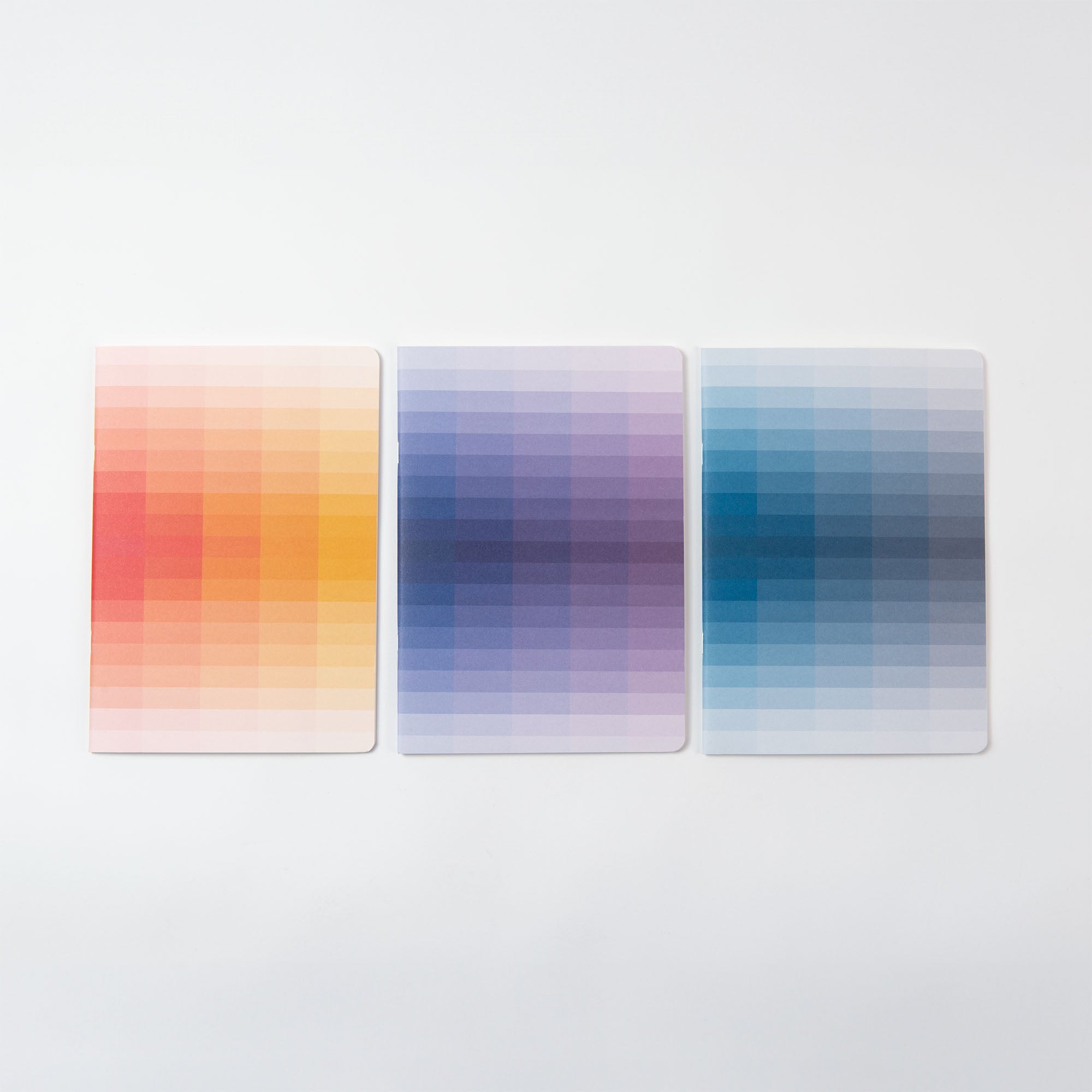 Drei Notizhefte der Desktop Edition von paperlux.store liegen nebeneinander und zeigen ein von einem Designer entworfenes Pixelmuster mit Farbverlauf: das linke in Gelb- und Rottönen, das mittlere in Lilatönen und das rechte in Blau- und Blaugrüntönen vor einem schlichten weißen Hintergrund.