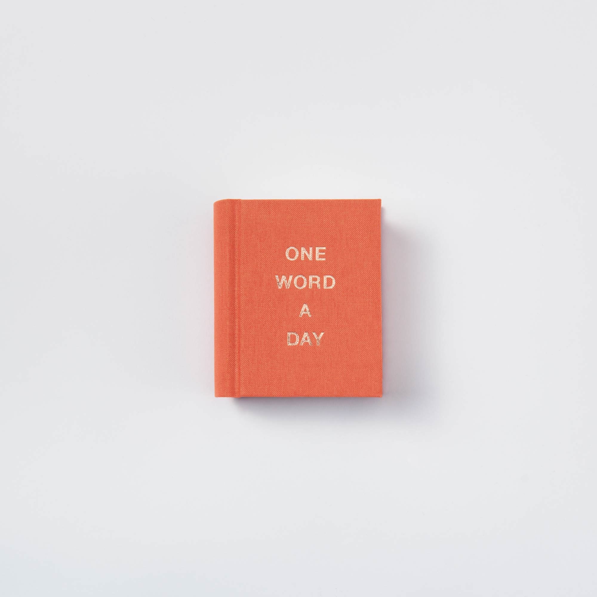 Ein kleines, geschlossenes, orangefarbenes OWAD Elements Mini-Tagebuch von paperlux mit "ONE WORD A DAY" in weißer Schrift auf dem Einband, handgefertigt in Hamburg - ein einzigartiges Geschenk, platziert auf einem schlichten weißen Hintergrund.