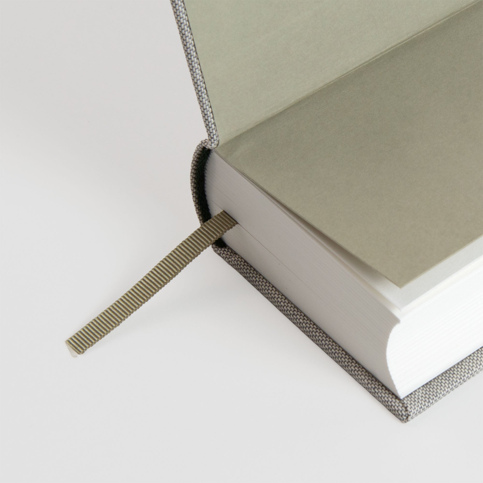 Eine Nahaufnahme eines aufgeschlagenen Hardcover-Buches der OWAD Hygge Collection von paperlux.store mit einem strukturierten grauen Einband. Zwischen den Seiten befindet sich ein gestreiftes Lesezeichen aus Stoff, und das Buch liegt auf einer weißen Oberfläche.