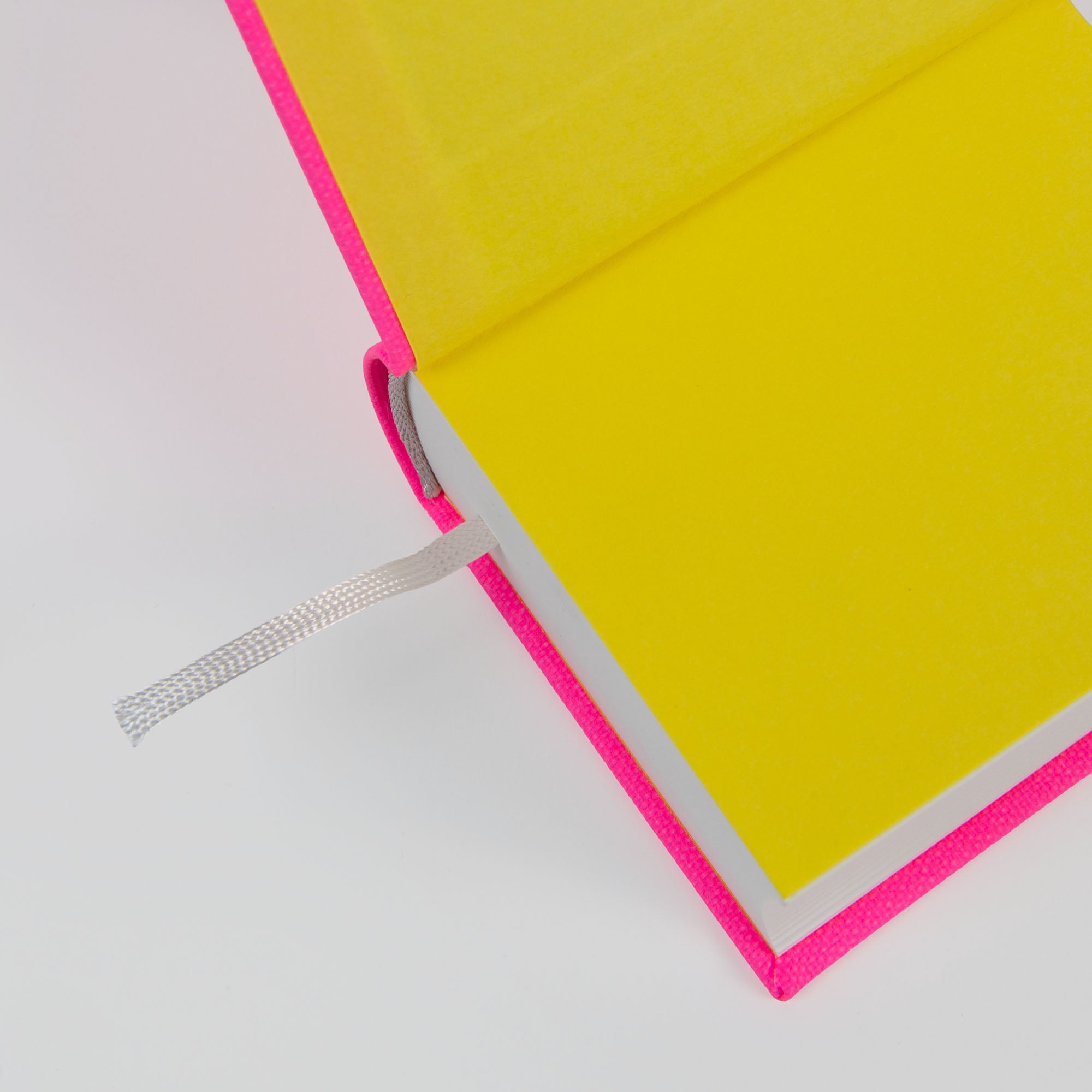 Ein aufgeschlagenes paperlux OWAD Neon Notizbuch mit leuchtend gelben Seiten, einem pinkfarbenen Einband und einem silbernen Lesezeichen ist von oben auf einer weißen Fläche fotografiert - ideal für Tagebucheinträge oder als kreative Geschenkidee.
