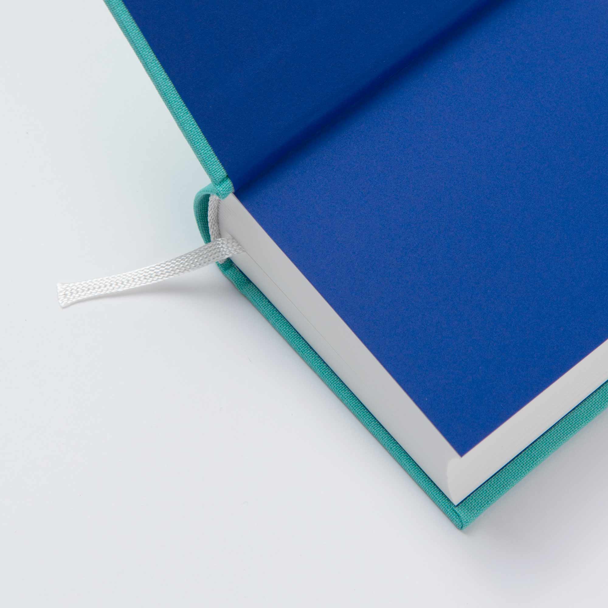 Nahaufnahme eines aufgeschlagenen OWAD Shine Bright-Hardcovers von paperlux mit einem blaugrünen Einband, blauem Vorsatzpapier, weißen Seiten und einem Lesezeichen mit weißem Band - perfekt als Mini-Notizbuch oder nachdenkliches Geschenk -, das flach auf einer weißen Oberfläche liegt.