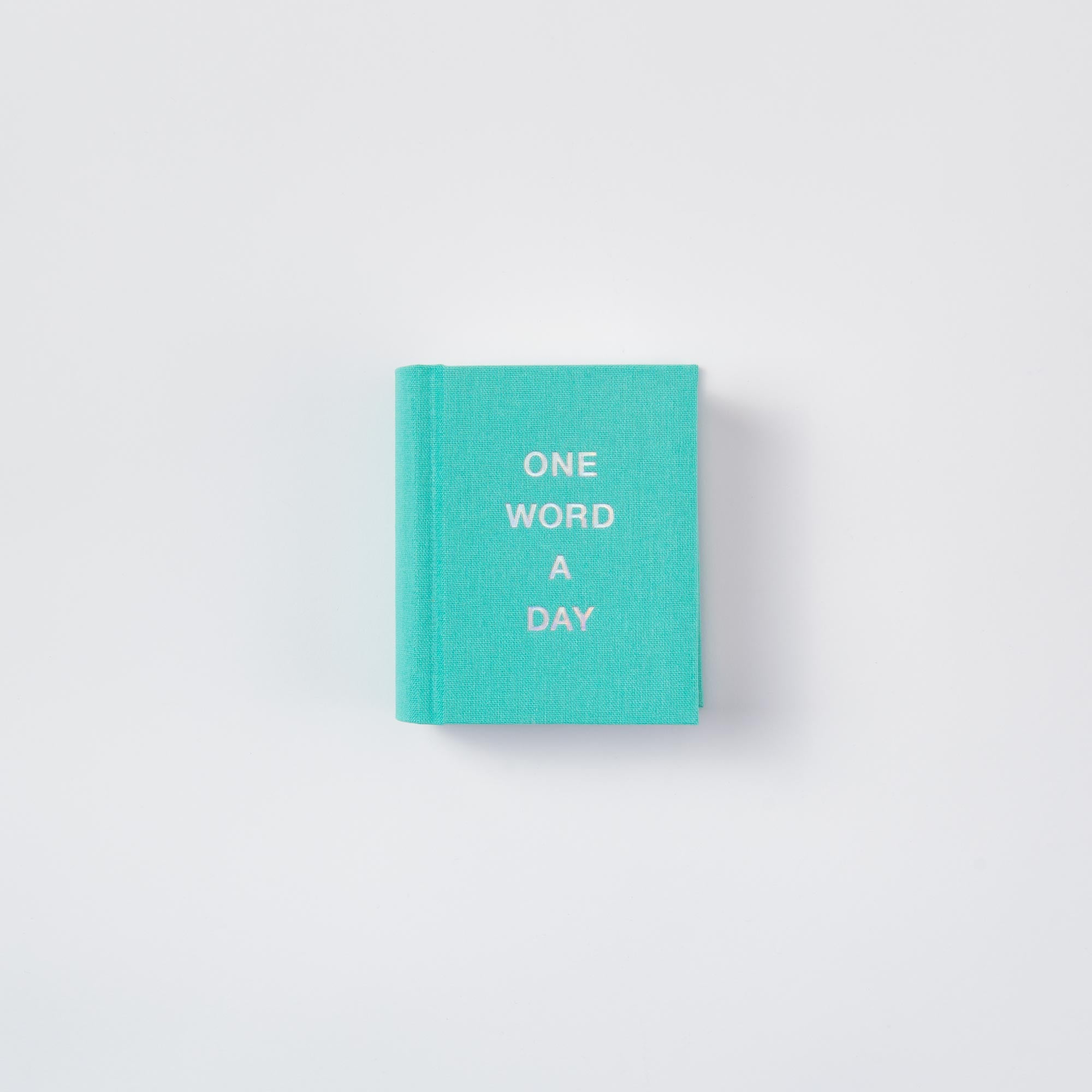 Ein kleines türkisfarbenes Mini-Notizbuch aus Papierlux mit dem Namen OWAD Shine Bright, mit "ONE WORD A DAY" in weißer Schrift auf dem Einband und auf einem schlichten weißen Hintergrund - ideal als nachdenkliches Geschenk.