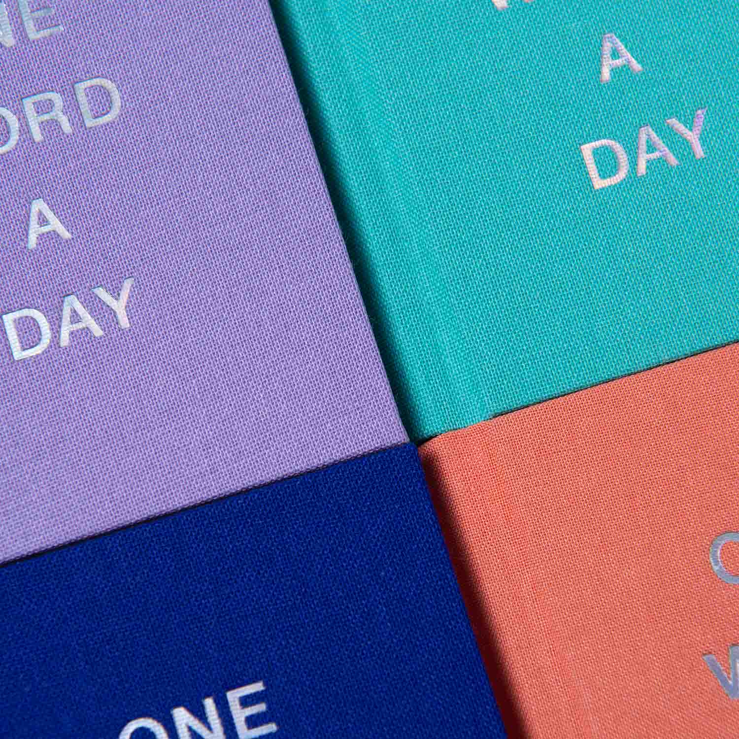 Nahaufnahme von vier farbenfrohen Buchdeckeln - lila, blaugrün, marineblau und korallenrot - aus der OWAD Shine Bright-Serie von paperlux. Jedes dieser Mini-Notizbücher ist mit dem fettgedruckten weißen Text "One Word a Day" versehen und damit ein ideales Geschenk für Schreibwarenliebhaber.