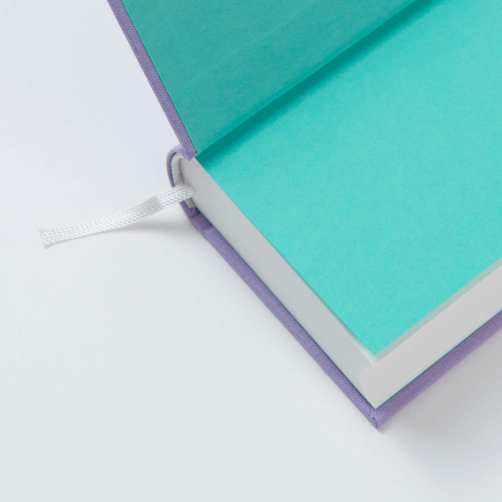 Ein aufgeschlagenes OWAD Shine Bright Hardcover von paperlux mit lilafarbenem Einband, türkisem Vorsatzpapier und weißem Lesezeichen liegt auf einer weißen Fläche - perfekt als Geschenk oder als persönliches One Word a Day Mini-Notizbuch.