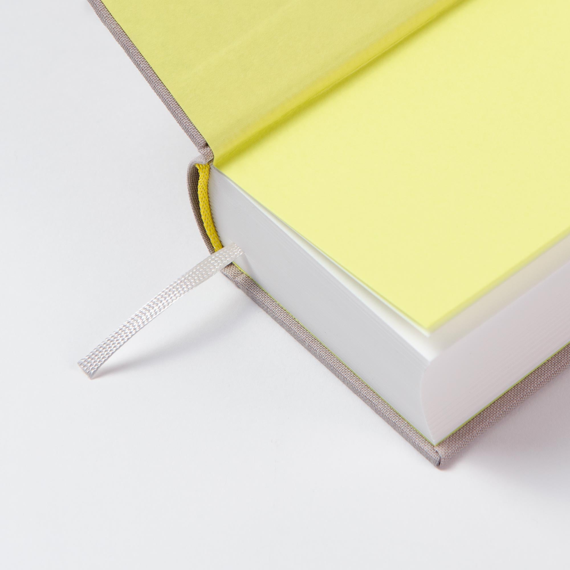Eine Nahaufnahme der Ecke eines geöffneten, handgefertigten OWAD Urban Leinen Notizbuches von paperlux mit gelbem Einband, grauem Stofflesezeichen und Vorsatzblättern - ideal als Geschenk Hochzeit - auf einer weißen Fläche ruhend.