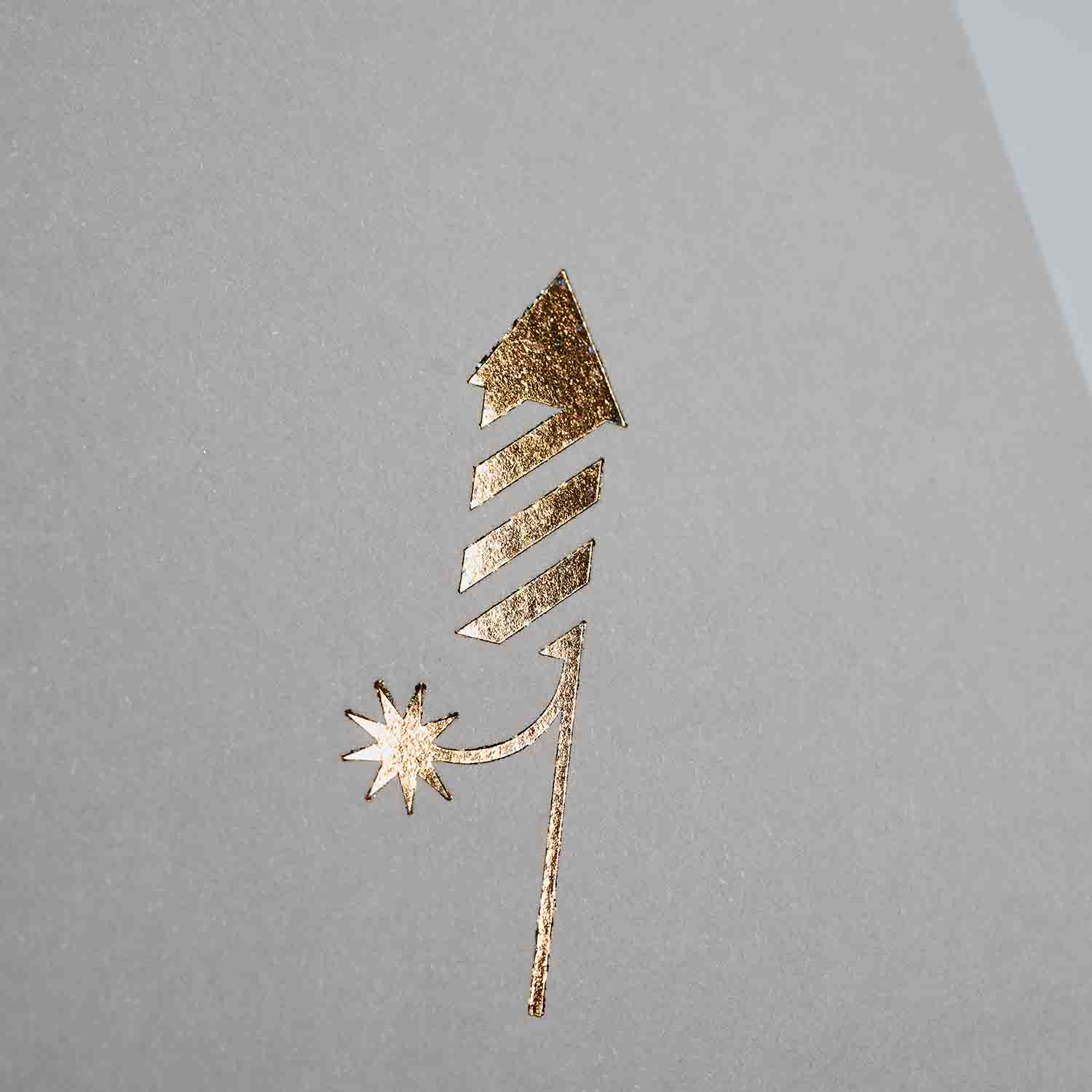 Eine goldgeprägte Feuerwerksrakete mit gestreiftem Körper und sternförmigem Funken leuchtet auf hellgrauem Hintergrund - perfekt als einzigartige Klappkarte oder im Weihnachtskarten 3-er Set Madarine, Silber, Schokolade von paperlux.store.