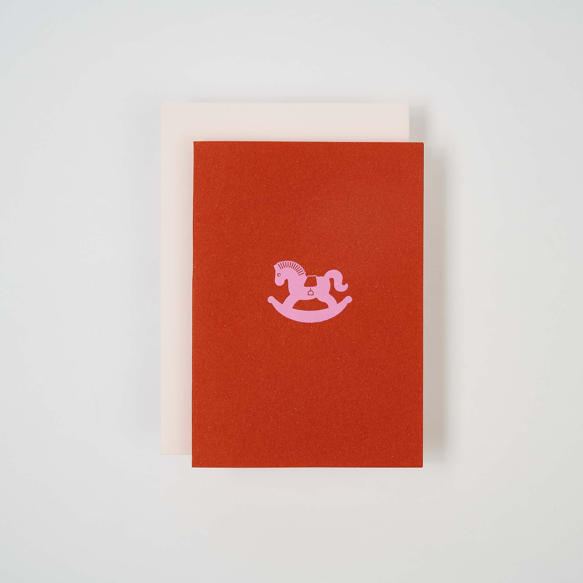 Auf weißem Untergrund präsentieren zwei geschlossene Notizbücher ihr minimalistisches Design. Das obere, einer Geburtstagskarte von paperlux.store ähnelnd, zeigt die Karte Schaukelpferd mit rotem Cover und rosa Illustration. Darunter befindet sich ein schlichtes beiges Notizbuch; beide eignen sich ideal für Ihre Klappkartensammlung.