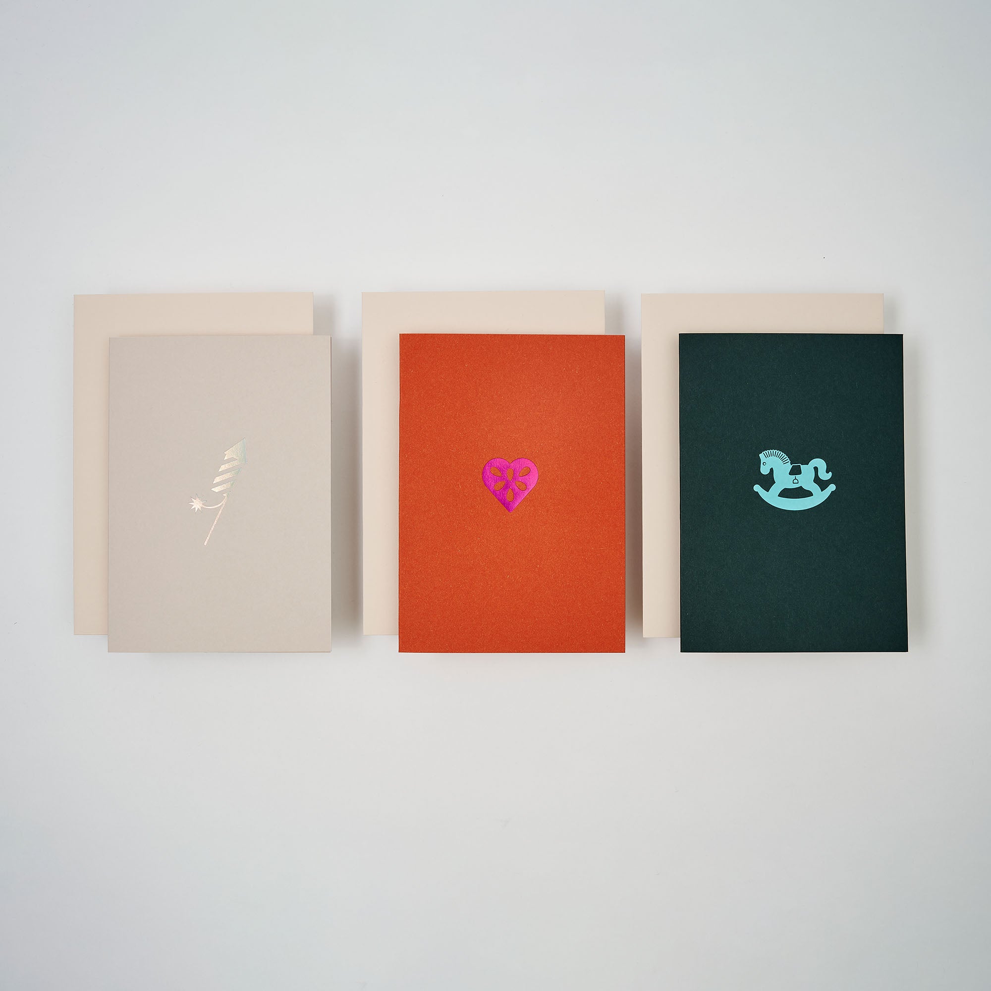 Das Weihnachtskarten 3er-Set von paperlux.store enthält drei A6-Karten: beige mit goldener Feder, orange mit rosa Herz und Blume und dunkelgrün mit blauem Schaukelpferd. Jede Karte wird mit einem passenden Umschlag geliefert.