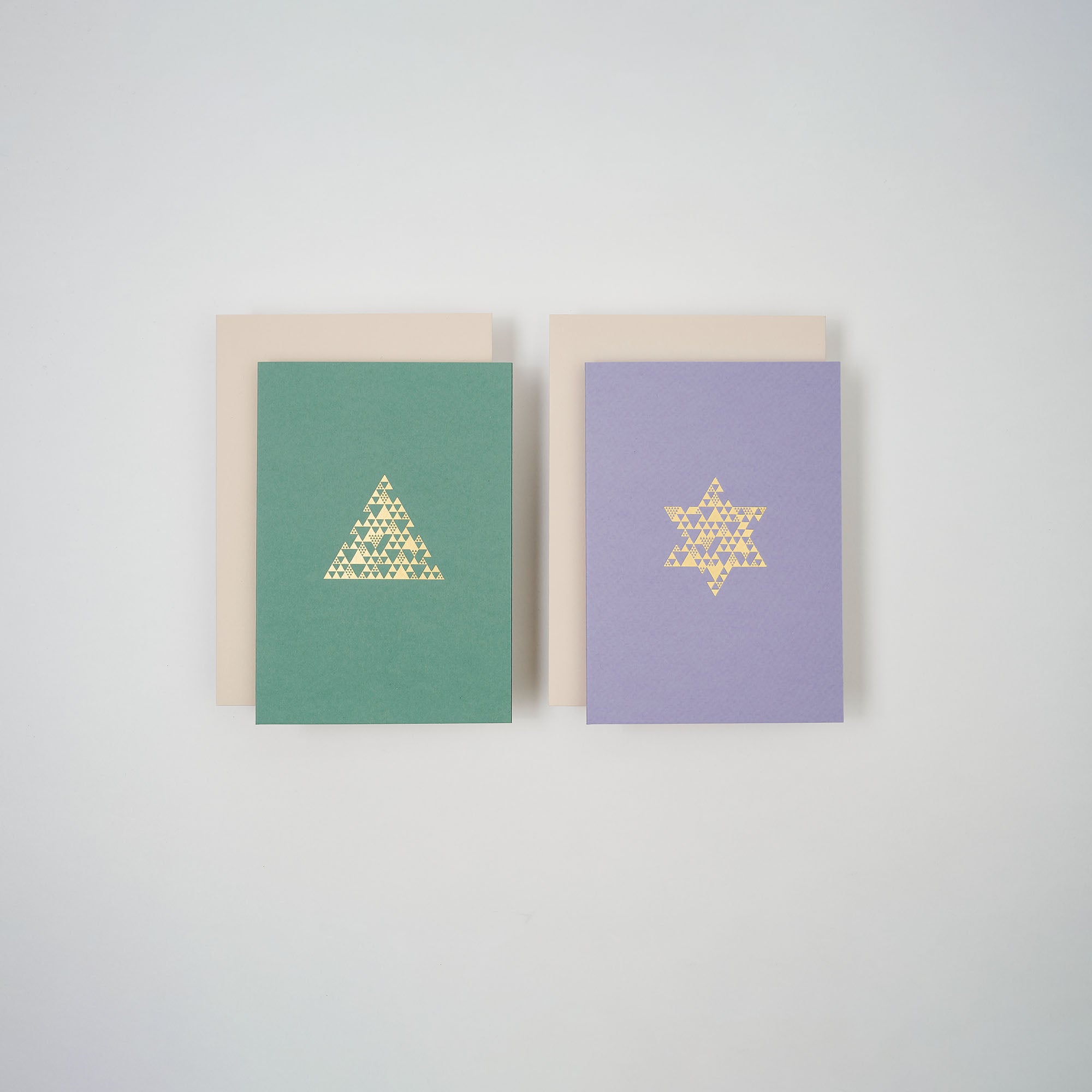 Zwei Karten aus dem paperlux.store Weihnachtskarten 2er-Set Lavendel & Salbei sind auf weißem Hintergrund abgebildet. Die linke Karte ist grün mit einem goldenen Dreieck, die rechte ist lila mit einem goldenen Davidstern. Hinter jeder Karte befinden sich beige Umschläge.