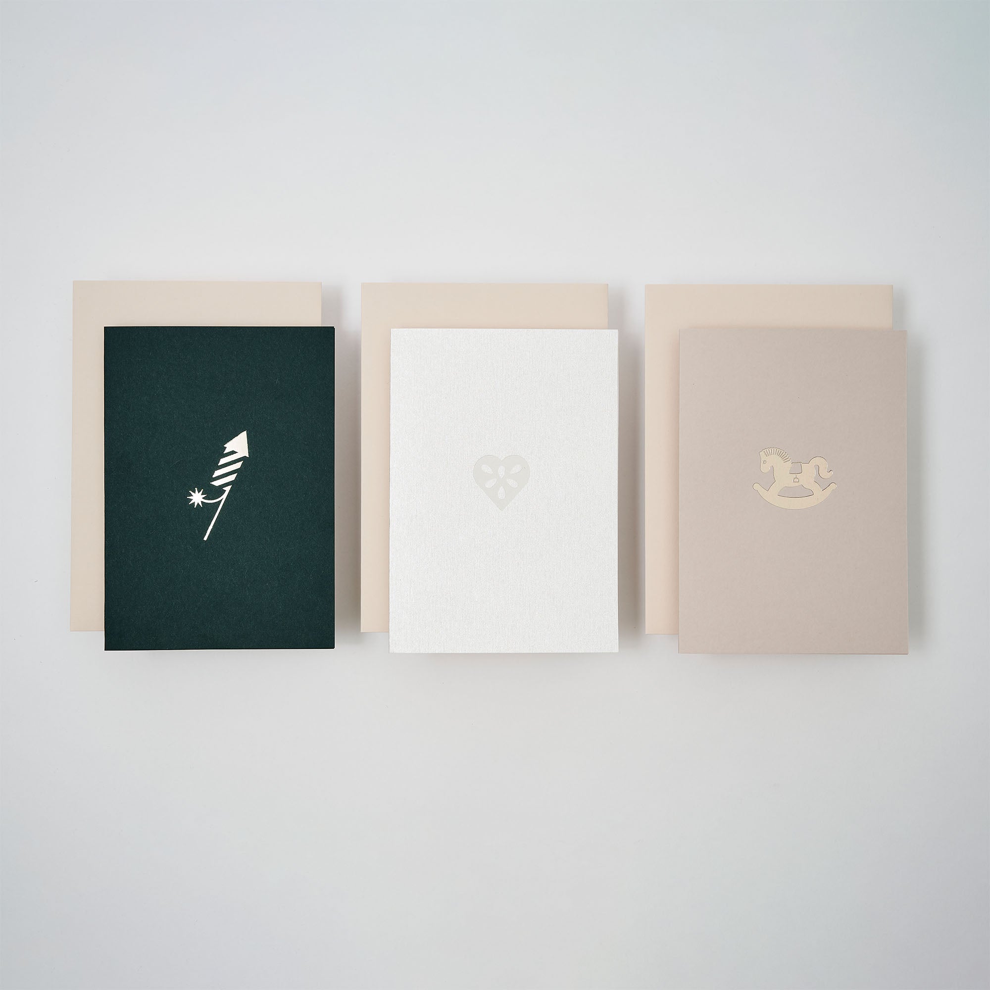 Das Weihnachtskarten 3er-Set Forest Green, Frost, Silver Dust von paperlux.store ist mit drei A6-Karten und Umschlägen abgebildet: eine Feder mit Stern auf dunkel, ein Herz auf weiß und ein Schaukelpferd auf beige - jeweils in minimalistischem Design.