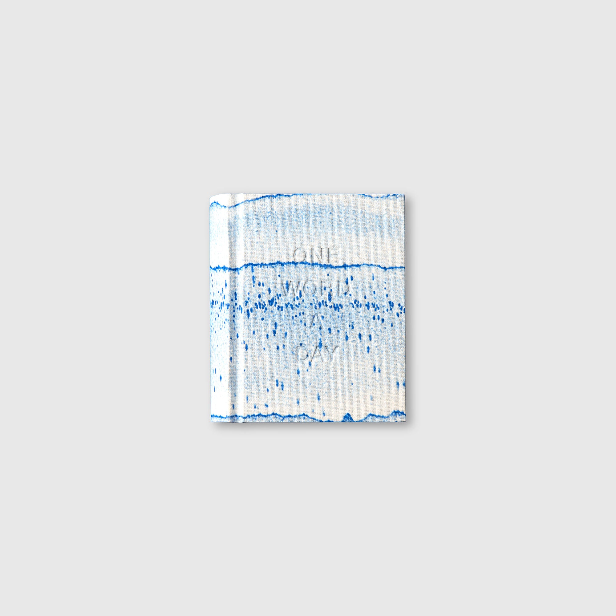 Die OWAD x TORTUS - Limited Edition von paperlux.store ist ein kleines Buch mit blau-weißem, abstraktem Aquarell-Cover, hellgrauem Hintergrund und dezent geprägtem ONE WORD A DAY-Text, inspiriert von Keramik-Handwerk.