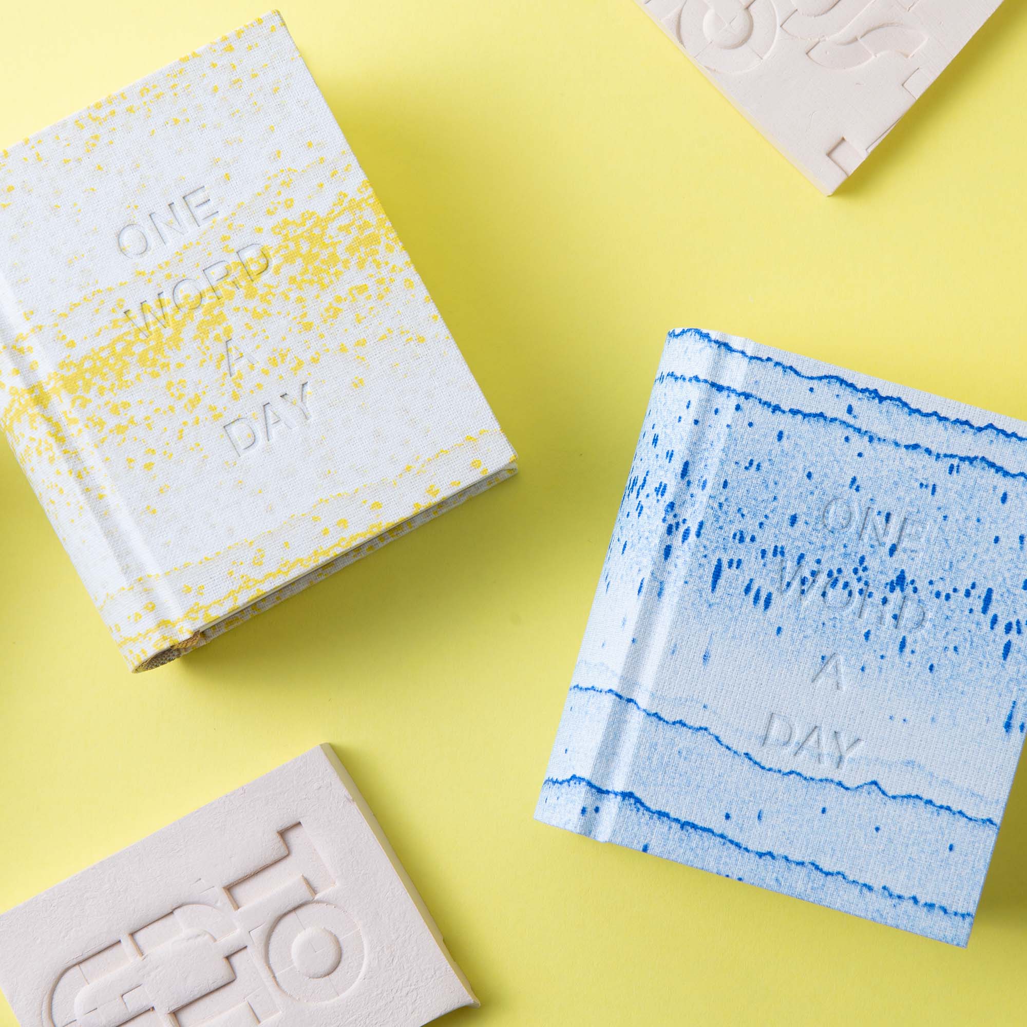 Zwei strukturierte OWAD x TORTUS - Limited Edition Bücher von paperlux.store in Gelb und Blau ruhen auf einem pastellgelben Hintergrund, daneben zwei rechteckige, geprägte Tonstücke, die an das Keramik-Handwerk erinnern.