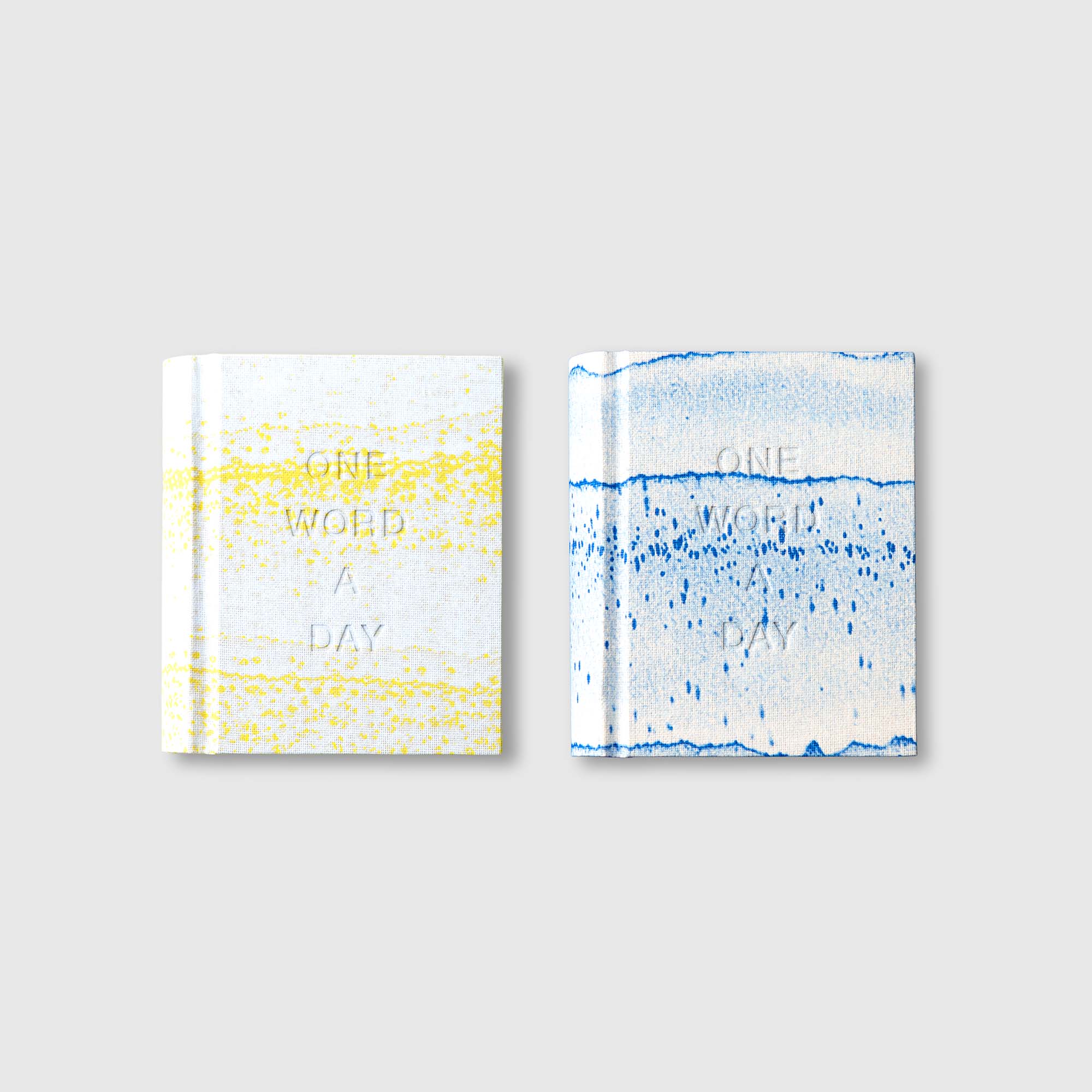 Zwei Bücher aus der OWAD x TORTUS - Limited Edition von paperlux.store mit abstrakten gelb-weißen und blau-weißen Einbänden und geprägten One Word a Day-Titeln werden nebeneinander auf einem hellgrauen Hintergrund präsentiert.