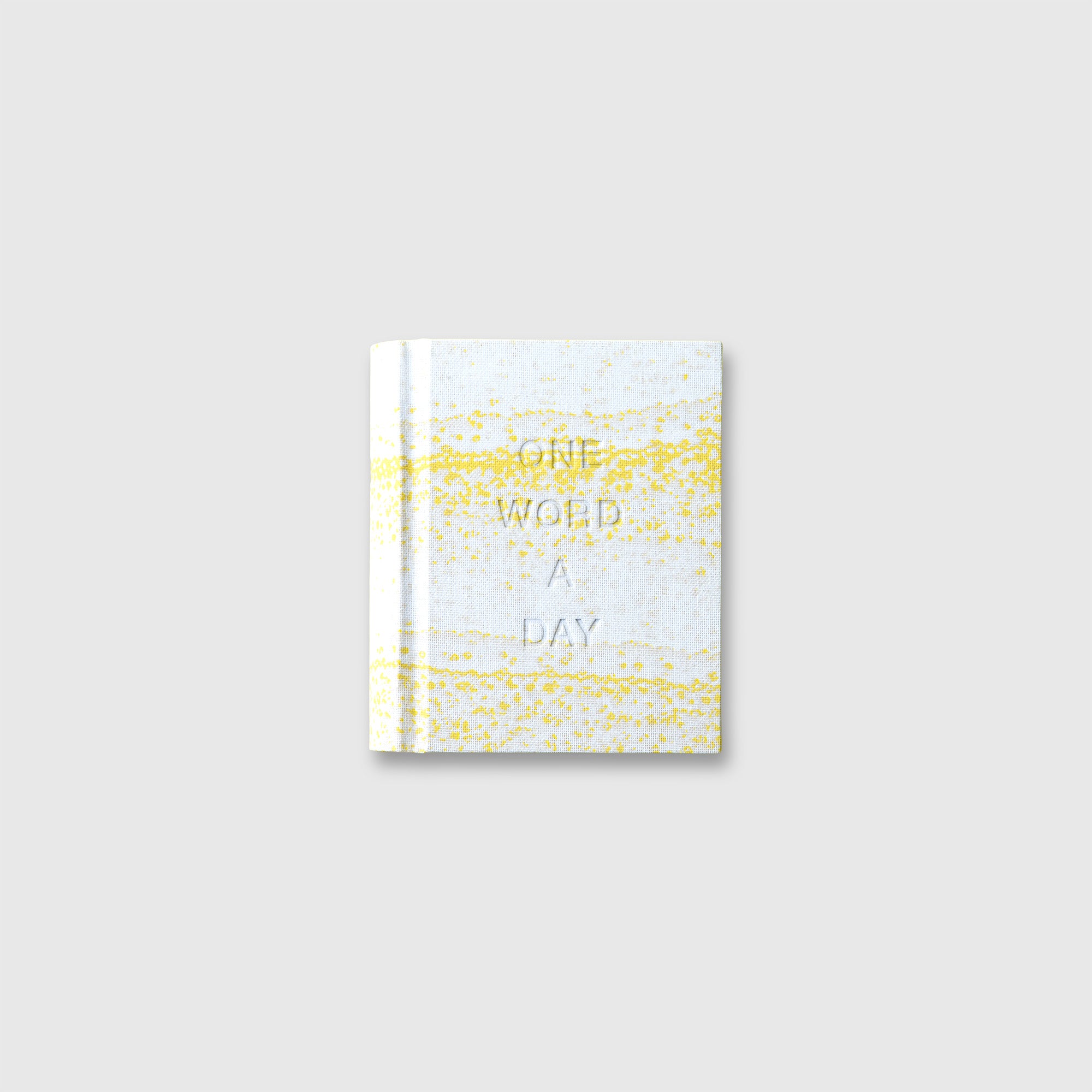 Ein OWAD x TORTUS Buch in limitierter Auflage von paperlux.store mit einem kleinen weißen Einband mit gelben Sprenkeln und geprägtem ONE WORD A DAY Titel, inspiriert von TORTUS Kopenhagens Keramik-Handwerk, dargestellt auf einem schlichten hellgrauen Hintergrund.