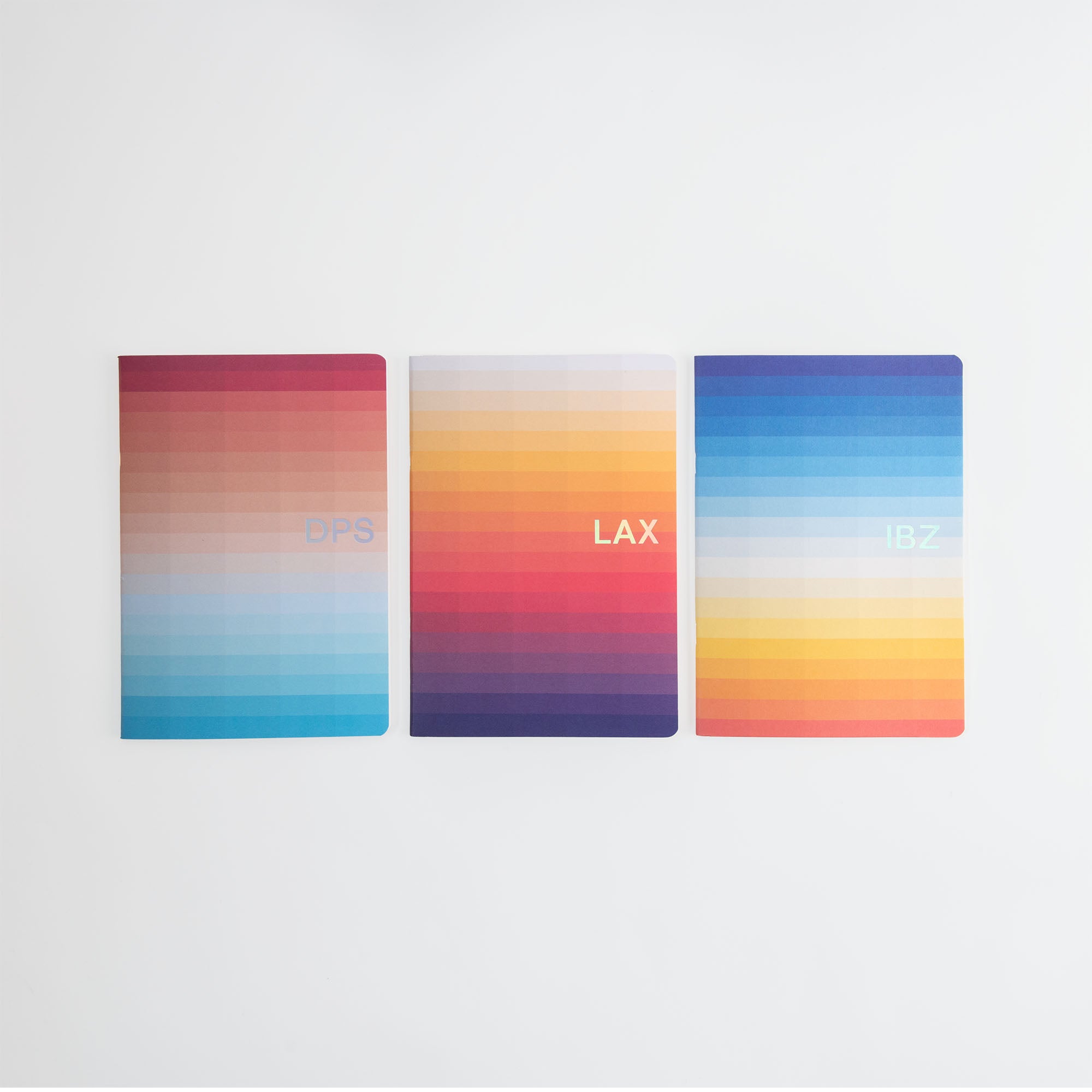 Die Sunset Edition Notizhefte A5 von paperlux.store umfasst drei Notizbücher mit Farbverlaufscovern in leuchtenden, sonnenuntergangsinspirierten Farbtönen wie Rot, Orange, Gelb, Blau und Lila. Jedes Notizbuch zeigt elegant den Flughafencode „DPS“, „LAX“ oder „IBZ“ auf einem strahlend weißen Hintergrund.