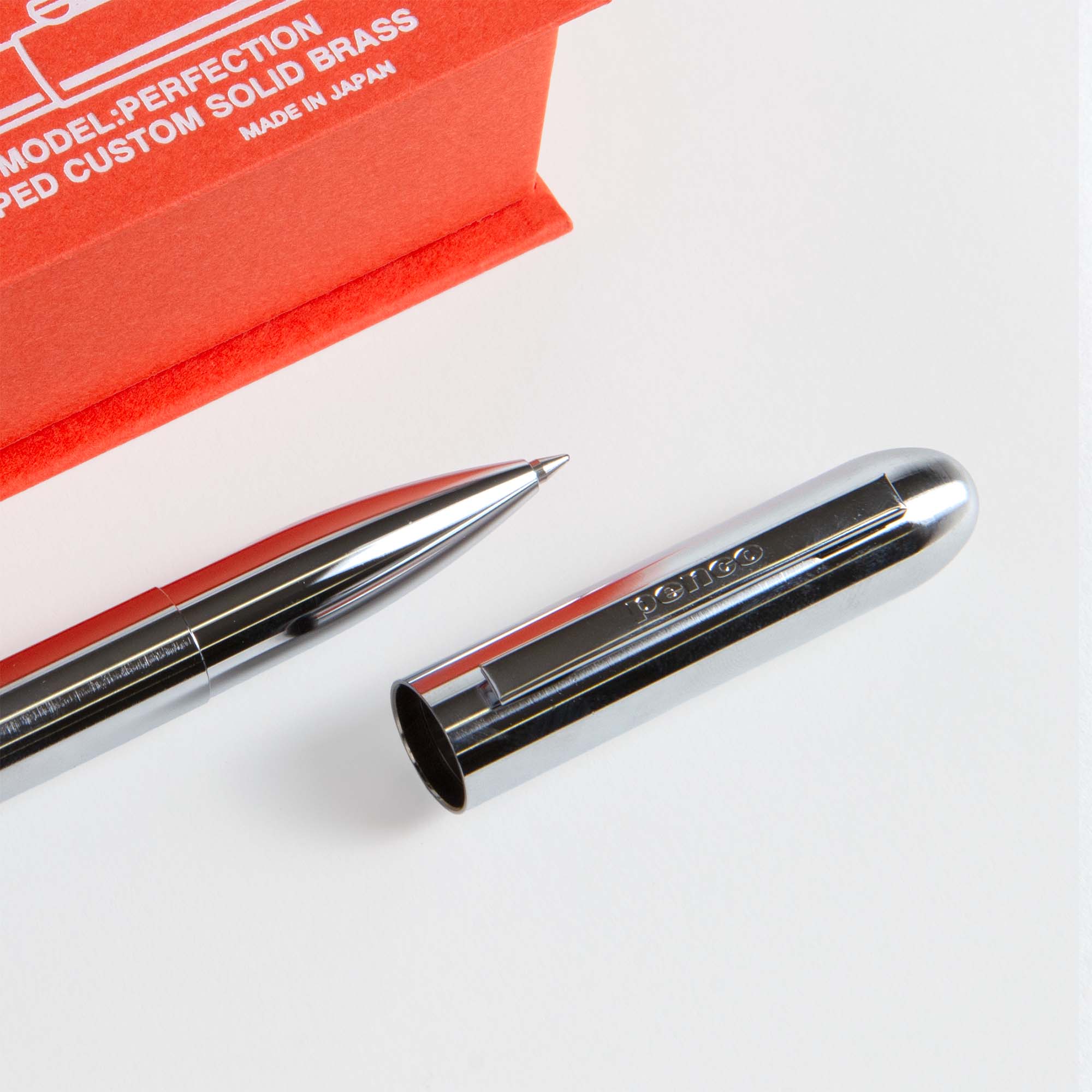 Ein Kompakt-Kugelschreiber - Bullet Pen von paperlux.store mit schwarzem Messinggehäuse liegt unverschlossen auf einer weißen Fläche neben einer roten Schachtel mit der Aufschrift "MODEL: PERFECTION" und "Hergestellt in Japan".