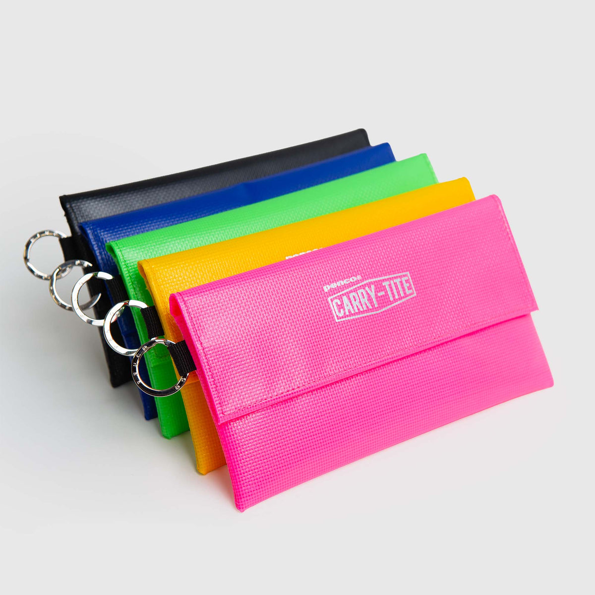 Fünf farbenfrohe Penco Carry Tite Tarp-Stiftetuis – mit Schlüsselringen in Rosa, Gelb, Grün, Blau und Schwarz – sind diagonal auf einem weißen Hintergrund abgebildet. Auf dem rosa Etui sticht der Schriftzug „CARRY-TITE“ hervor. Die von Penco aus robustem PVC-Planenstoff hergestellten Federmäppchen sind stilvolle und strapazierfähige, wasserabweisende Accessoires für den täglichen Gebrauch.