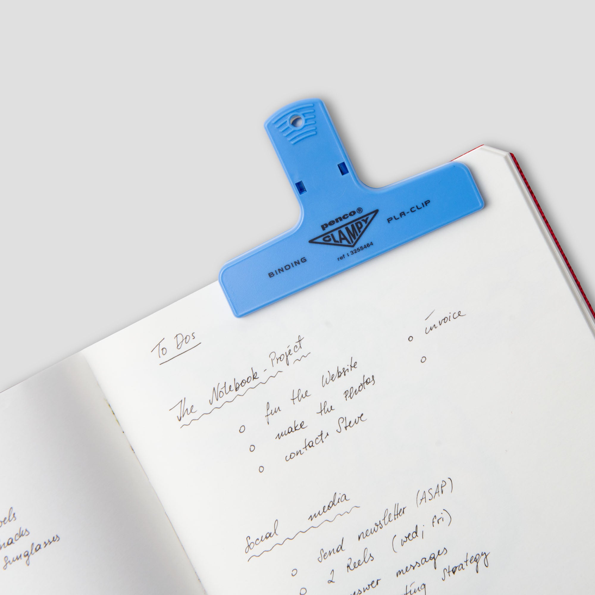 Ein offenes Notizbuch mit handgeschriebenen To-do-Listen, das oben mit einem großen blauen „Penco Clampy PlaClip“ Kunststoff von Penco zusammengehalten wird. Die Notizen enthalten Erinnerungen zum „Notebook-Projekt“ und zu den sozialen Medien.