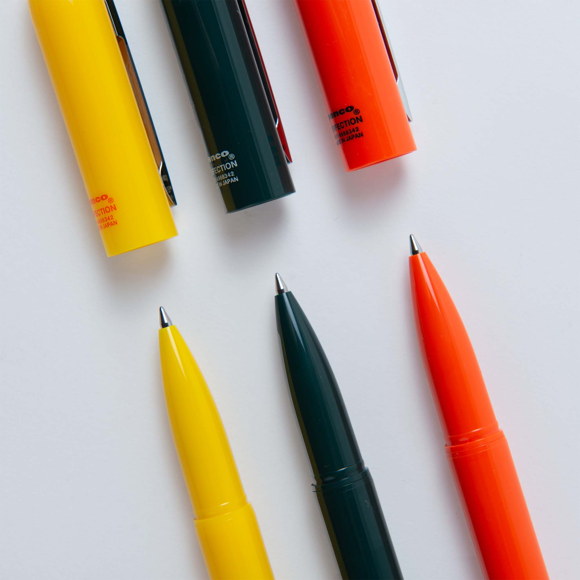 Drei Bullet-Kugelschreiber von Penco ohne Kappe sind diagonal angeordnet. Die eleganten gelben, schwarzen und orangefarbenen Gehäuse und Kappen mit glänzenden feinen Spitzen heben japanische Handwerkskunst hervor, da die Kappen parallel über ihren jeweiligen Stiften auf einem makellos weißen Hintergrund ruhen.