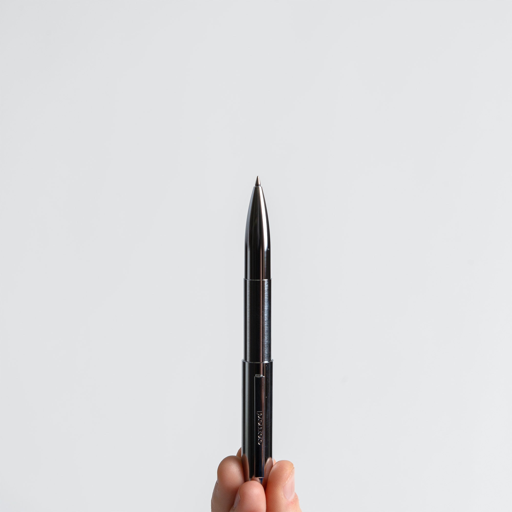 Eine Hand hält einen Kompakt-Kugelschreiber - Bullet Pen von paperlux.store, schwarzes Messing, made in Japan, aufrecht vor einem schlichten weißen Hintergrund.