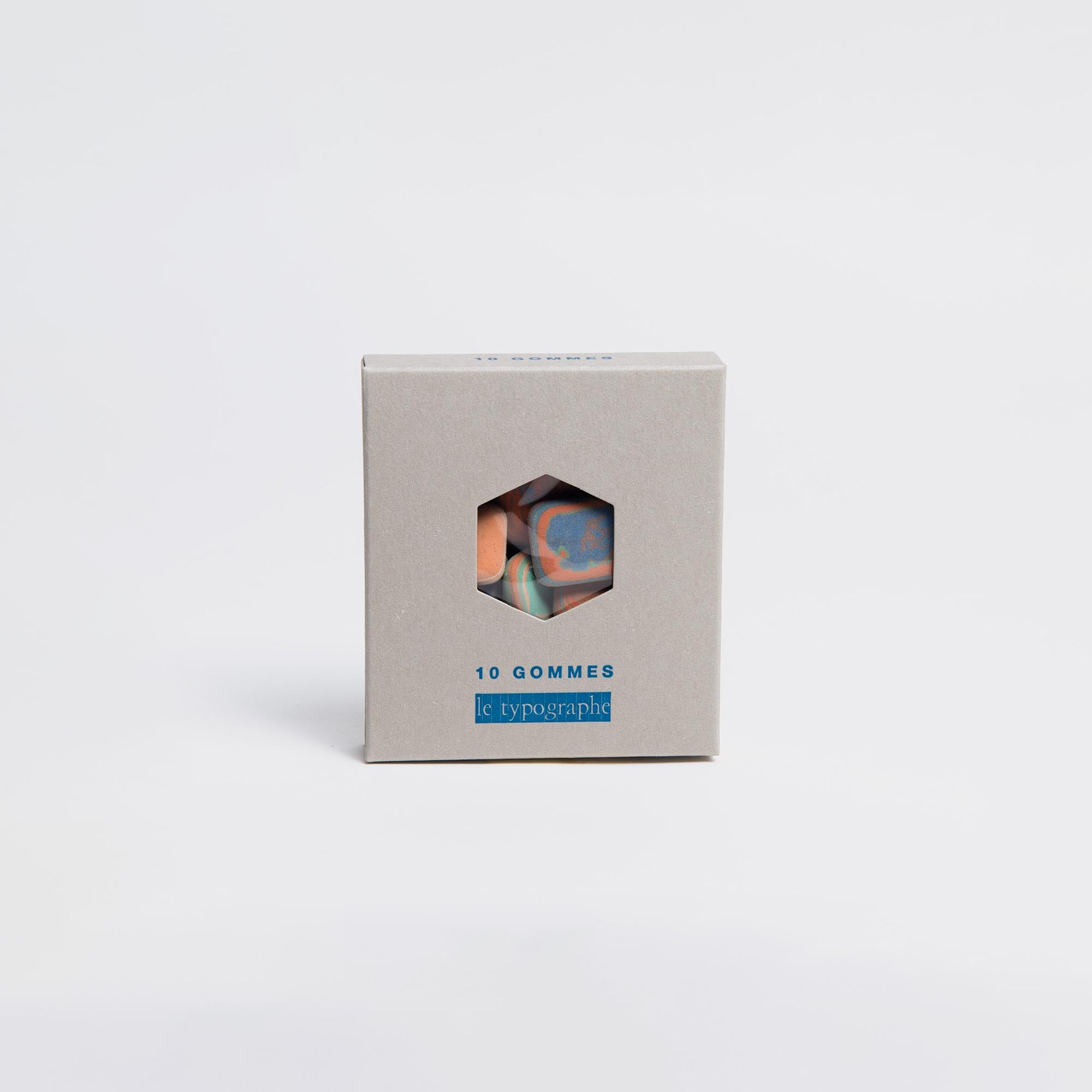 Eine Letterpress-Box aus Pappe mit der Aufschrift „10 Gommes“ und sechseckigem Ausschnitt zeigt bunte Radiergummis „Kieselsteine“. Auf der Box ist „Le Typographe“ in Blau auf minimalistischem weißen Hintergrund zu sehen.