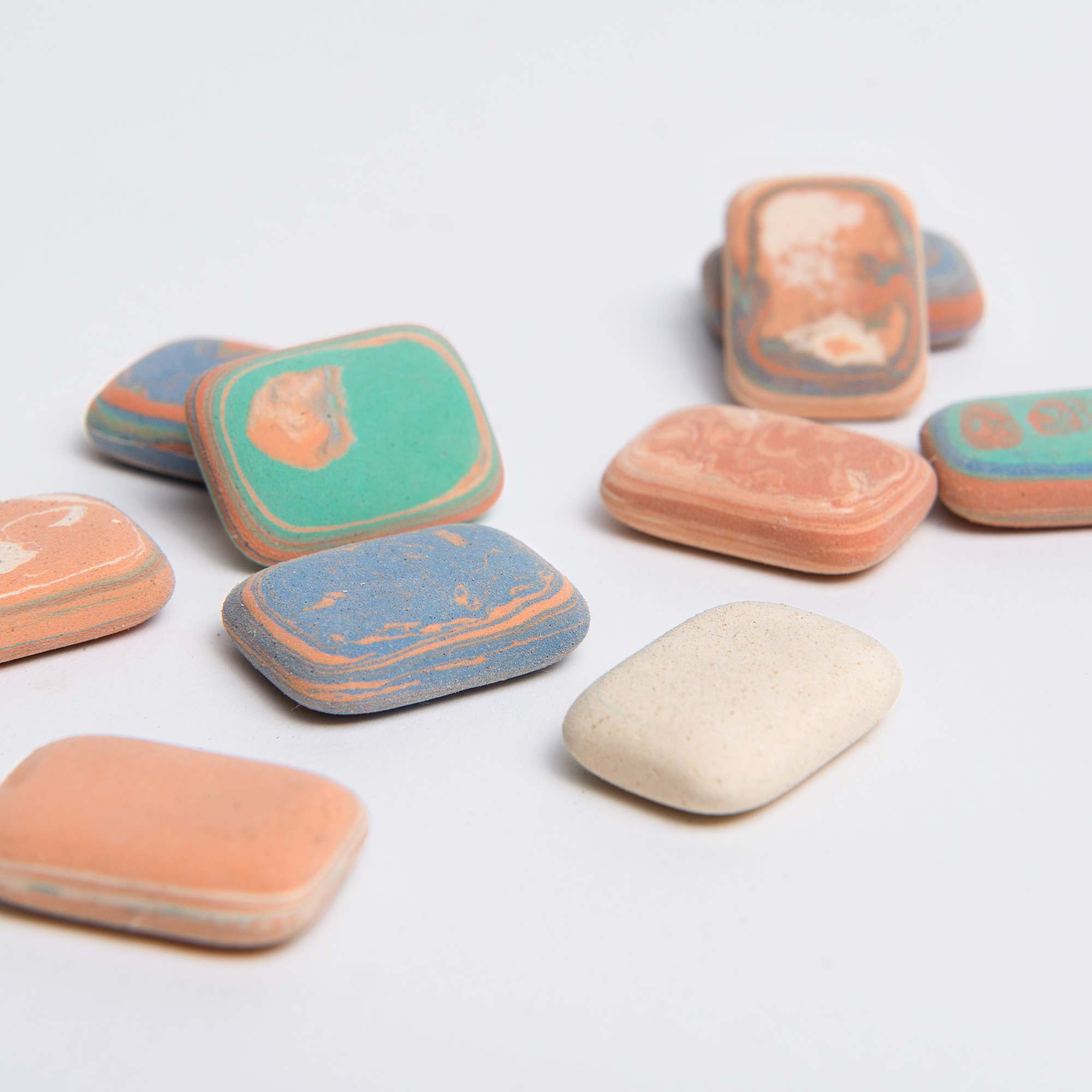 Eine Nahaufnahme der Radiergummis „Kieselsteine“ von Le Typographe, die auf einer weißen Oberfläche verstreut sind, zeigt rechteckige, mehrfarbige Radiergummis mit marmorierten Mustern in den Farbtönen Grün, Blau, Orange und Beige.