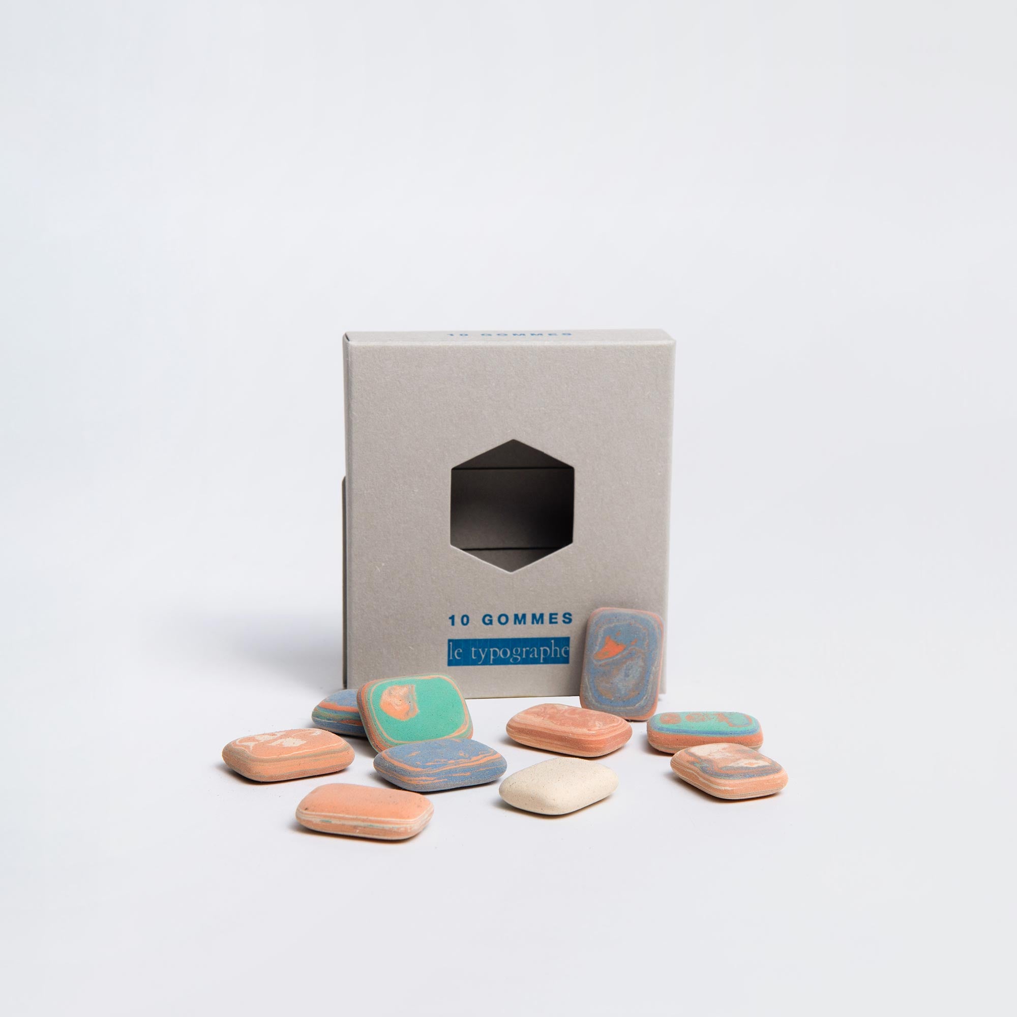 Eine Schachtel mit 10 Le Typographe Radiergummis „Kieselsteine“ ist geöffnet, vorne sind mehrere rechteckige Radiergummis in Orange, Blau und Weiß verstreut. Die hellbraune Buchdruckschachtel weist minimalen Text und einen geometrischen Ausschnitt auf.