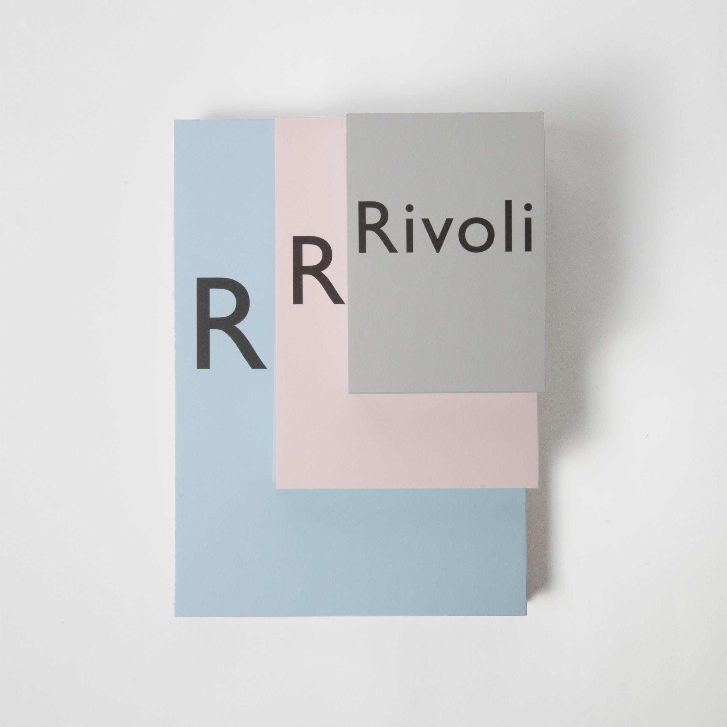 Drei gestapelte rechteckige Karten in Blau, Rosa und Grau aus hochwertigem Papier von Carta Pura. Jede Karte ist mit einem schwarzen „R“ versehen, die oberste graue Karte trägt die Aufschrift „Skizzen- und Schreibblock Rivoli hellgrau“. Das Design ist mehrschichtig und minimalistisch auf weißem Hintergrund.