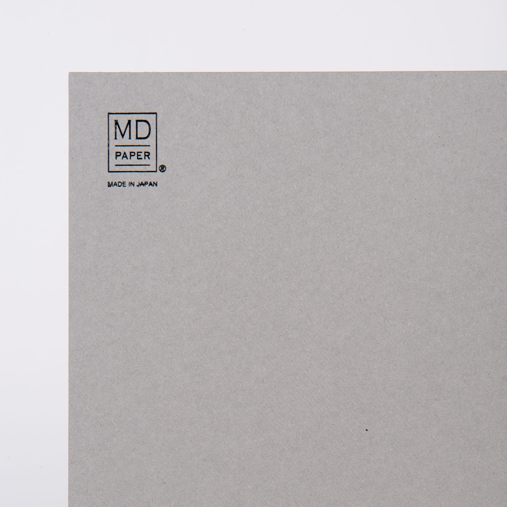 Nahaufnahme der oberen linken Ecke eines grauen Skizzenblocks A4 von Midori. „MD PAPER“ und „MADE IN JAPAN“ sind in Schwarz auf das minimalistische Design gedruckt, ideal für Notizen oder Skizzen, mit einem schlichten Hintergrund, auf dem nur die Marke zu sehen ist.