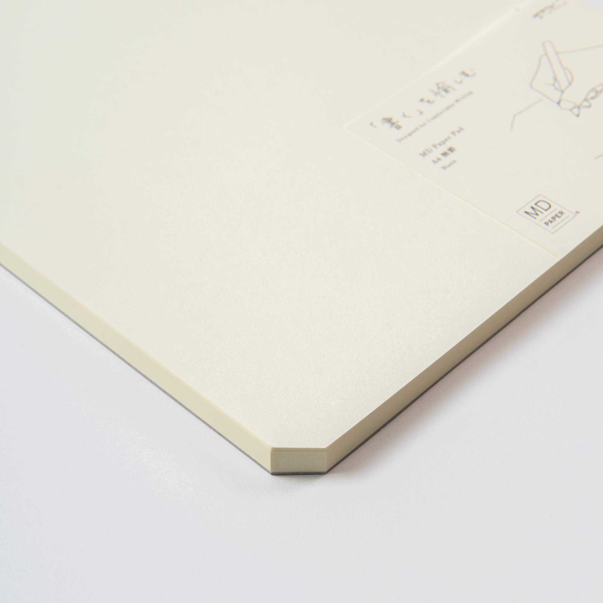 Ein beigefarbener Skizzenblock A4 von Midori, im minimalistischen Design mit japanischem Text und einer Stiftillustration, hebt das quadratische „MD“-Logo hervor. Dieses elegante Notizbuch ist perfekt für Skizzen oder Notizen und liegt auf einer hellen Oberfläche.