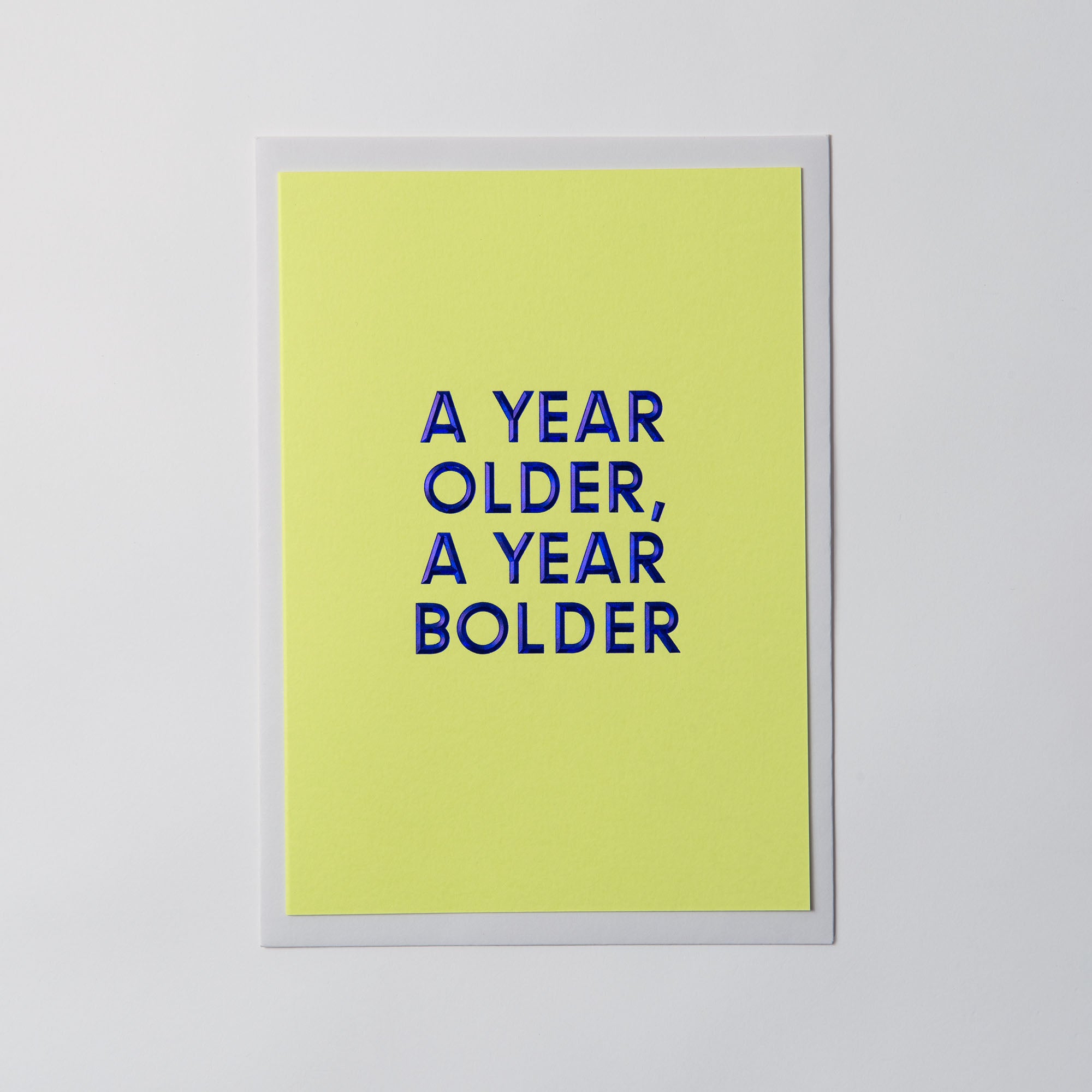 Die Geburtstagskarte „AYOAYB“ von paperlux.store zeigt „A YEAR OLDER, A YEAR BOLDER“ in kräftigem Blau auf leuchtend gelbem Hintergrund mit Heißfolienprägung, zentriert auf einer weißen Fläche.