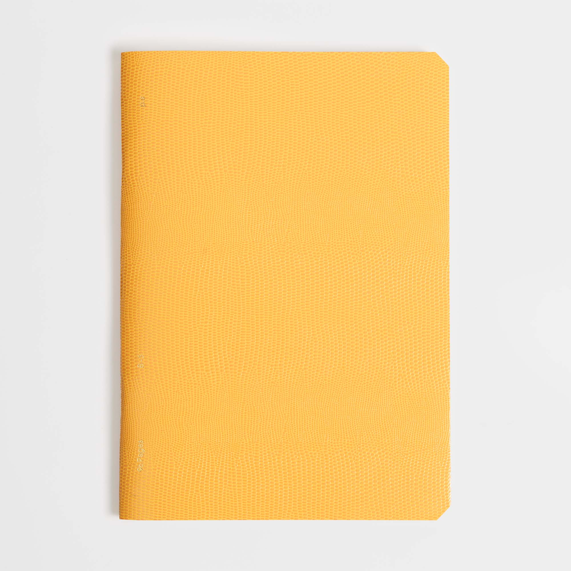 Ein geschlossenes gelbes Softcover-Notizbuch, das B96 Mellow Yellow von paperlux.store, mit strukturiertem Einband liegt auf einer weißen Fläche, vertikal ausgerichtet mit dem Rücken nach links.