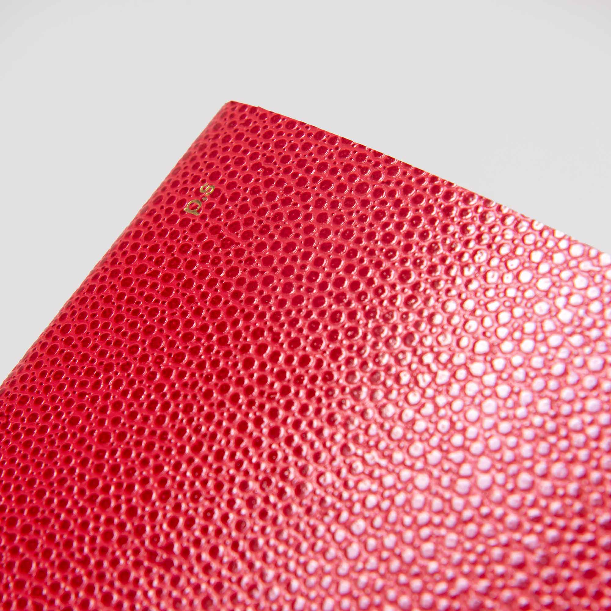 Nahaufnahme des paperlux.store B96 Red Lipstick Softcover-Notizbuchs aus strukturiertem rotem Kiesel-Leder mit winziger Goldprägung am Rand vor einem schlichten weißen Hintergrund.