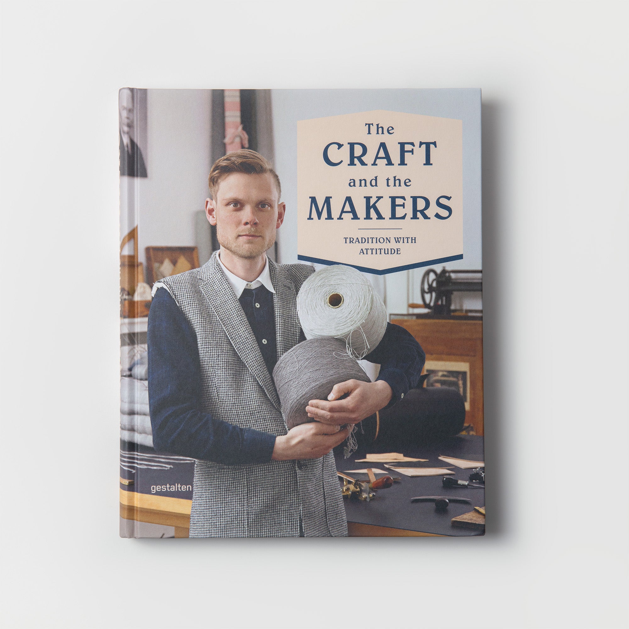 In „Das Handwerk und die Macher“ des Gestalten Verlags ist ein Mann im Anzug zu sehen, der große Garnrollen hält, vor der Kulisse eines inspirierenden Buchbinderei-Arbeitsplatzes voller Handwerkswerkzeuge und -materialien.
