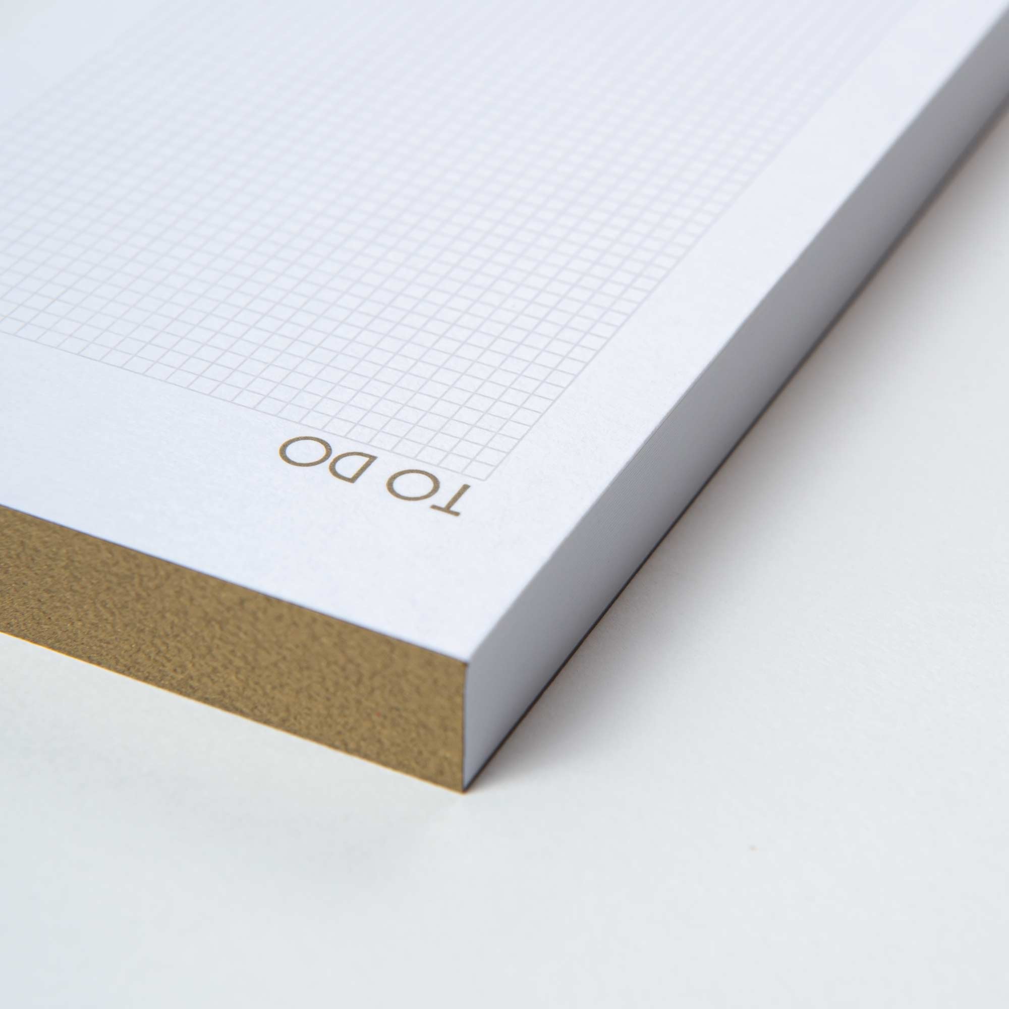 Nahaufnahme eines Notizblocks „To Do Block Gold“ von Le Typographe mit Gitterlinien auf weißem 300-g-Papier mit braunen Rändern. Handgefertigt in Brüssel, schräg auf einer schlichten weißen Oberfläche.