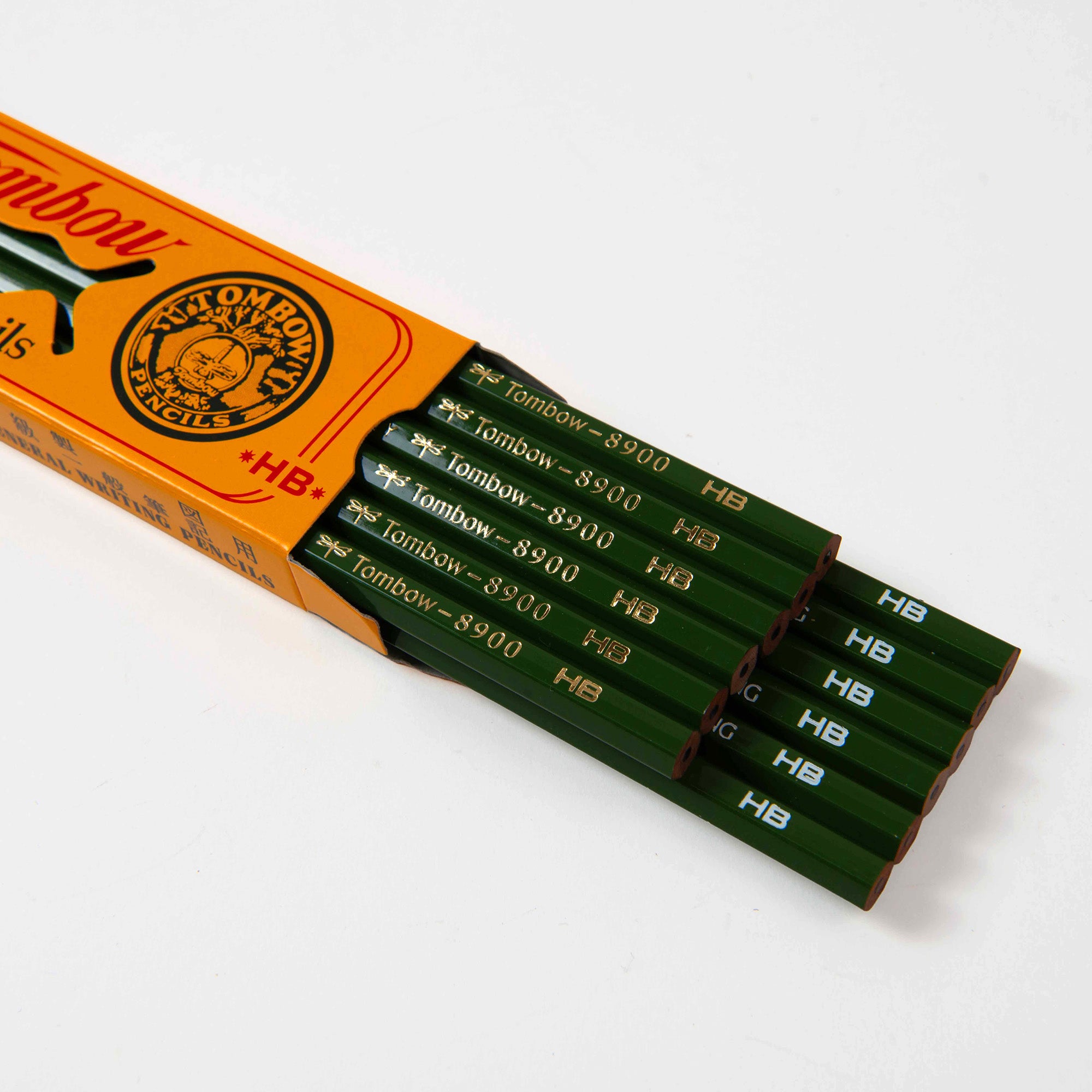 Eine gelbe 12er-Packung Tombow 8900 HB Bleistifte aus Japan, aus der einige grüne Bleistifte teilweise herausgeschoben wurden, trägt an den Seiten die goldene Aufschrift Tombow 8900 HB.