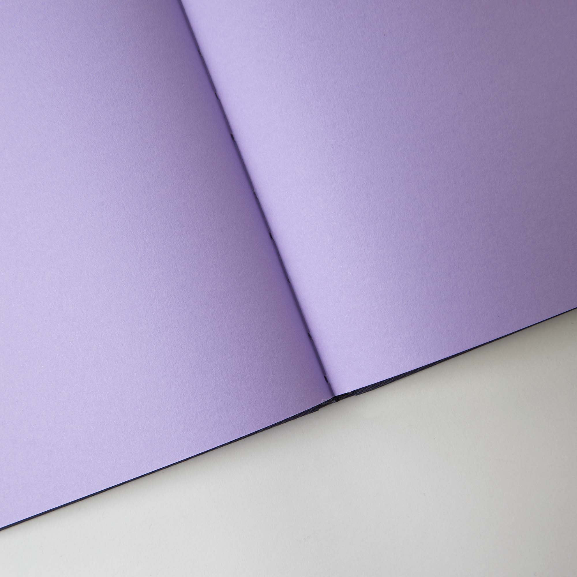 Ein violettes paperlux.store Notanizer Notizbuch A5 liegt offen auf einer weißen Oberfläche, seine makellosen Feinstpapierseiten werfen einen leichten Schatten entlang der Falte. Zeigen Sie mit jedem mit Farbschnitt verzierten Strich Kreativität und Stil – Sie sind Sie selbst.
