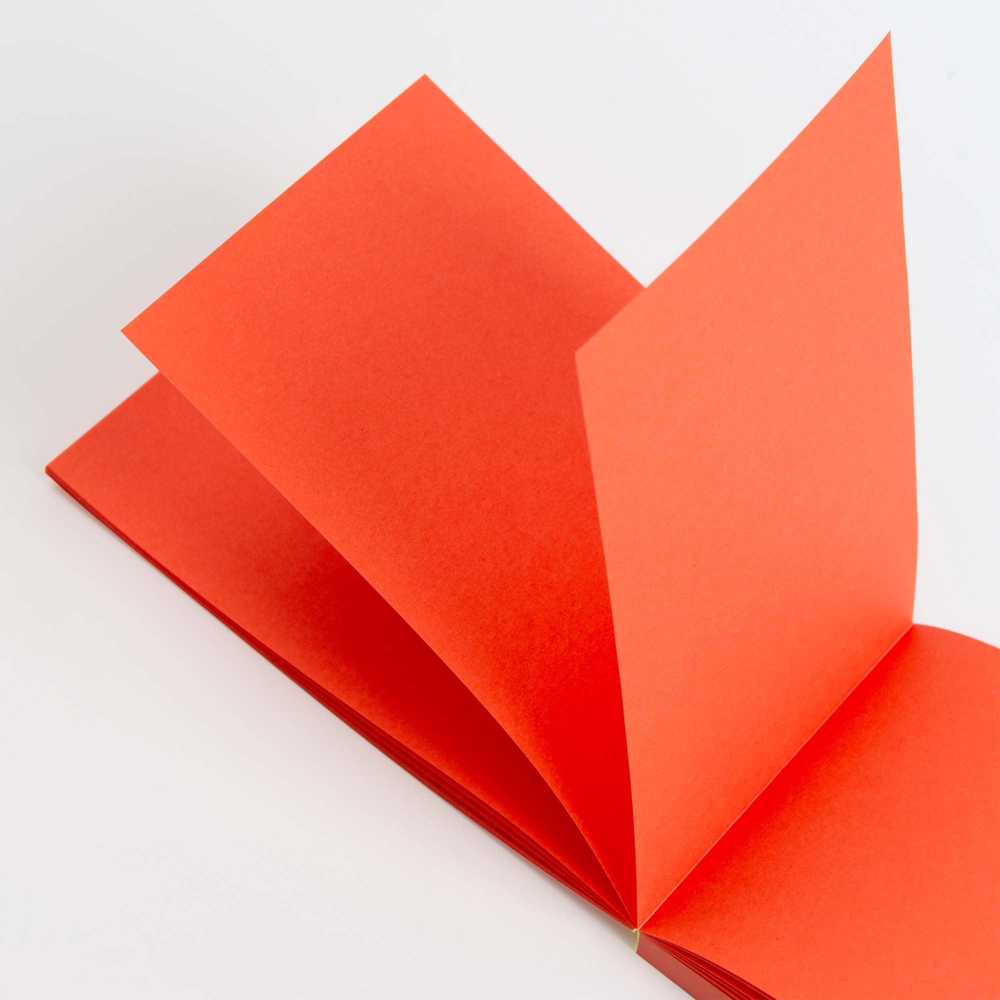 Nahaufnahme eines aufgeschlagenen paperlux Abreissblöcke Neon-Notizbuchs mit leuchtend roten Seiten aus Feinstpapier-Resten, von oben betrachtet auf weißem Hintergrund, wobei einige Seiten teilweise umgeschlagen sind, wodurch dynamische Winkel und Schatten entstehen.
