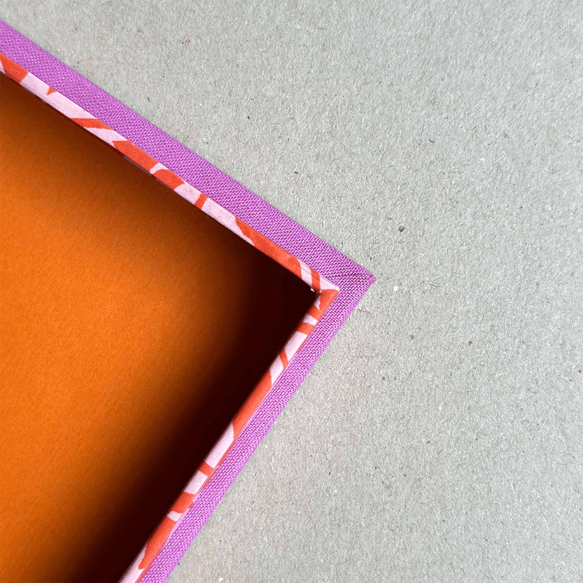 Eine Nahaufnahme der Ecke einer orangefarbenen rechteckigen Workshop Clamshell Box von paperlux.store mit rot-weiß gestreiften Rändern und einem rosa Rand auf einer neutralen Oberfläche - die perfekte Inspiration für Ihr nächstes Projekt.