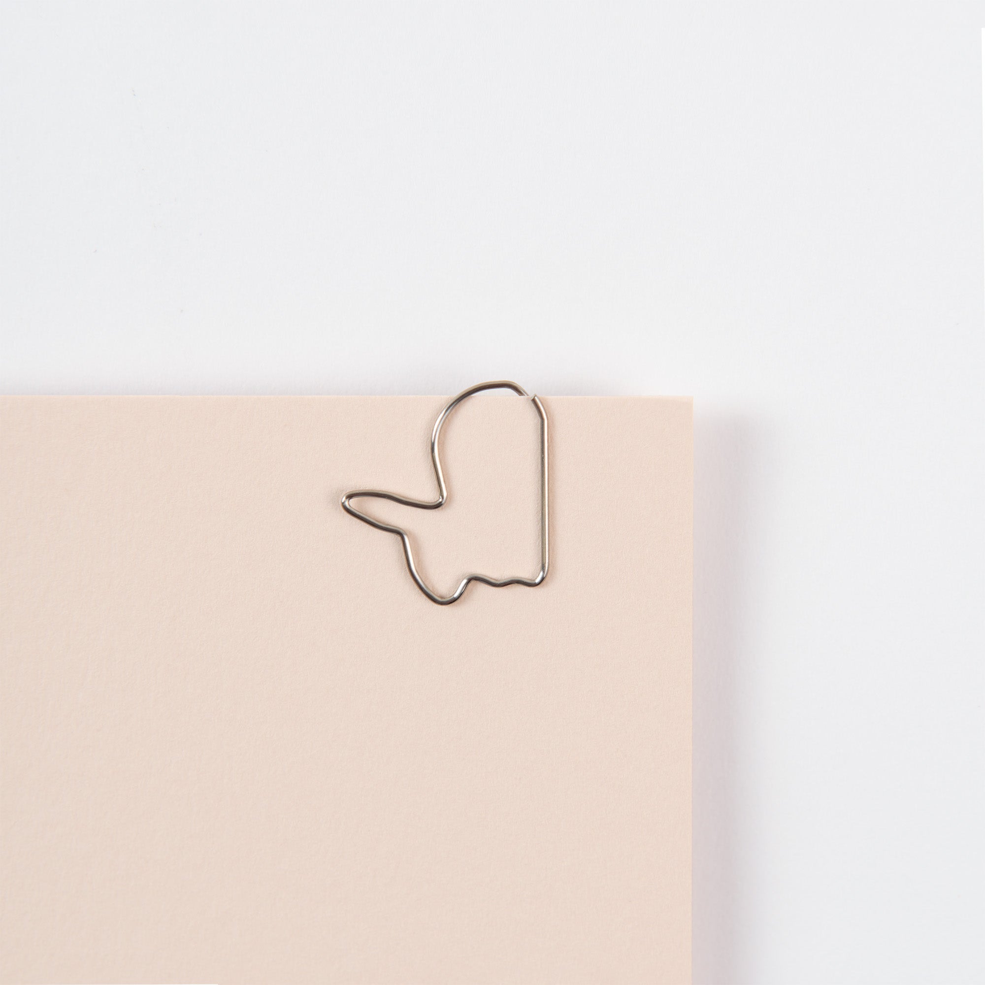 Ein Midori D-Clips: Büroklammern Hase in Hasenform ist vor einem schlichten weißen Hintergrund an der Ecke eines hellbeigen Blattes Papier befestigt.