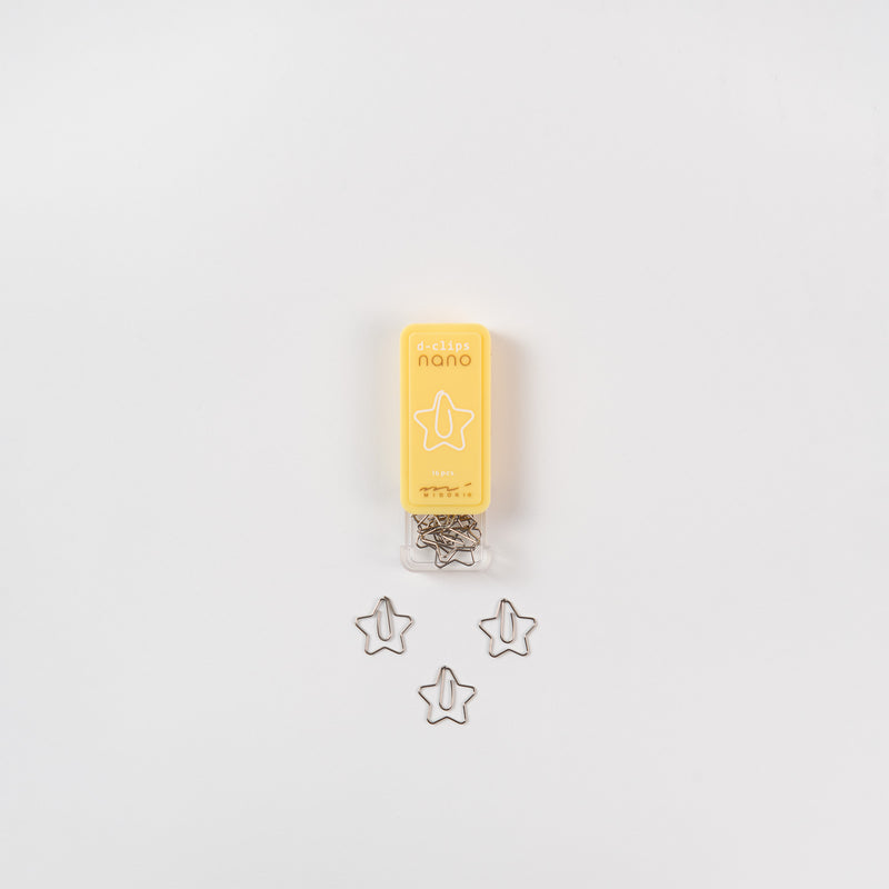 Eine geöffnete Schachtel mit sternförmigen Büroklammern liegt auf weißem Grund. Die gelbe Verpackung mit der Aufschrift „D-Clips Nano: Sterne“ von Midori eignet sich ideal, um Notiz- oder Liebesbriefen einen besonderen Charme zu verleihen. Unter der Schachtel sind drei Klammern übersichtlich angeordnet.